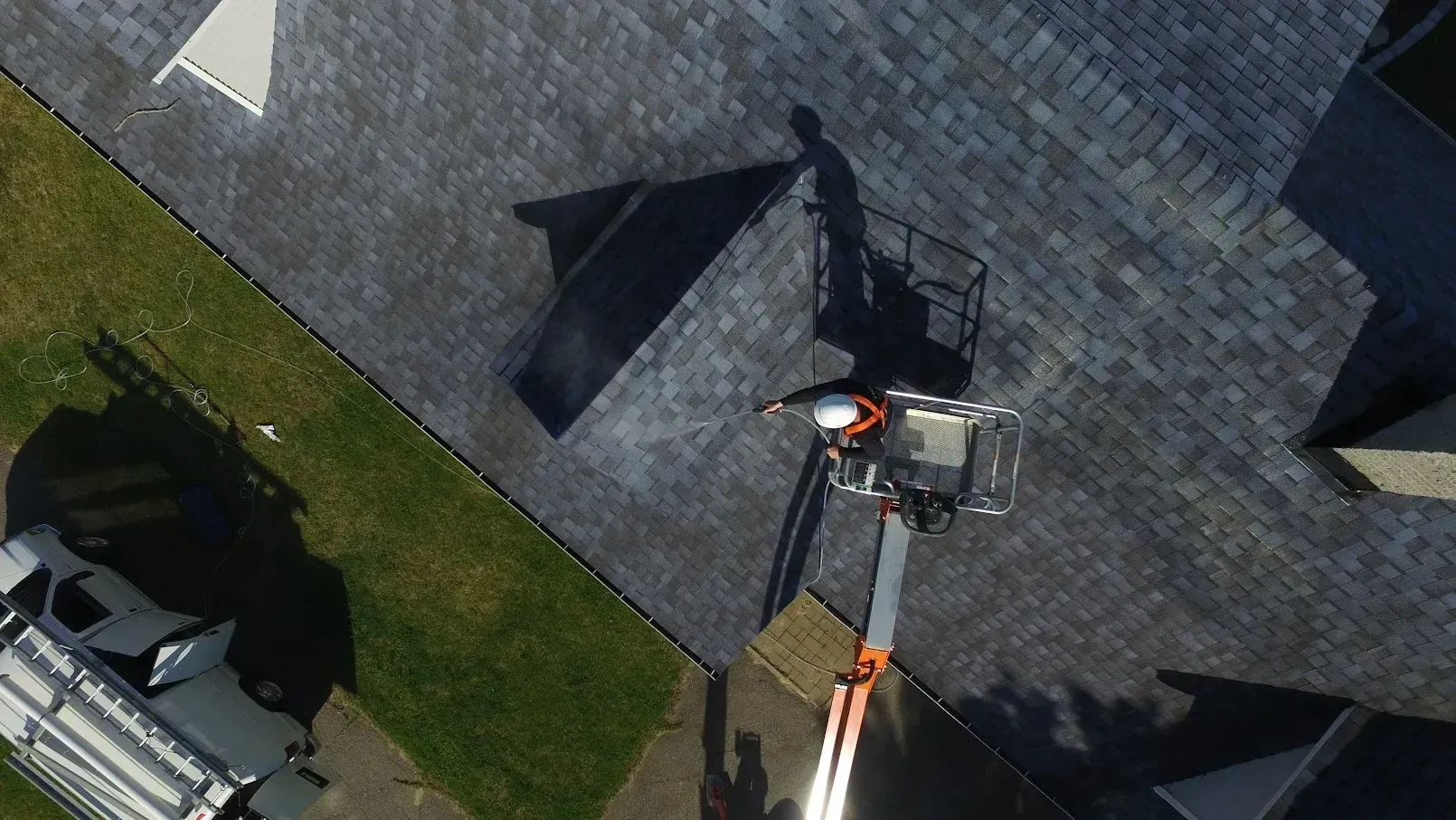 Aerial view of a person on a lift inspecting a gray shingle roof. A vehicle is on the ground nearby.