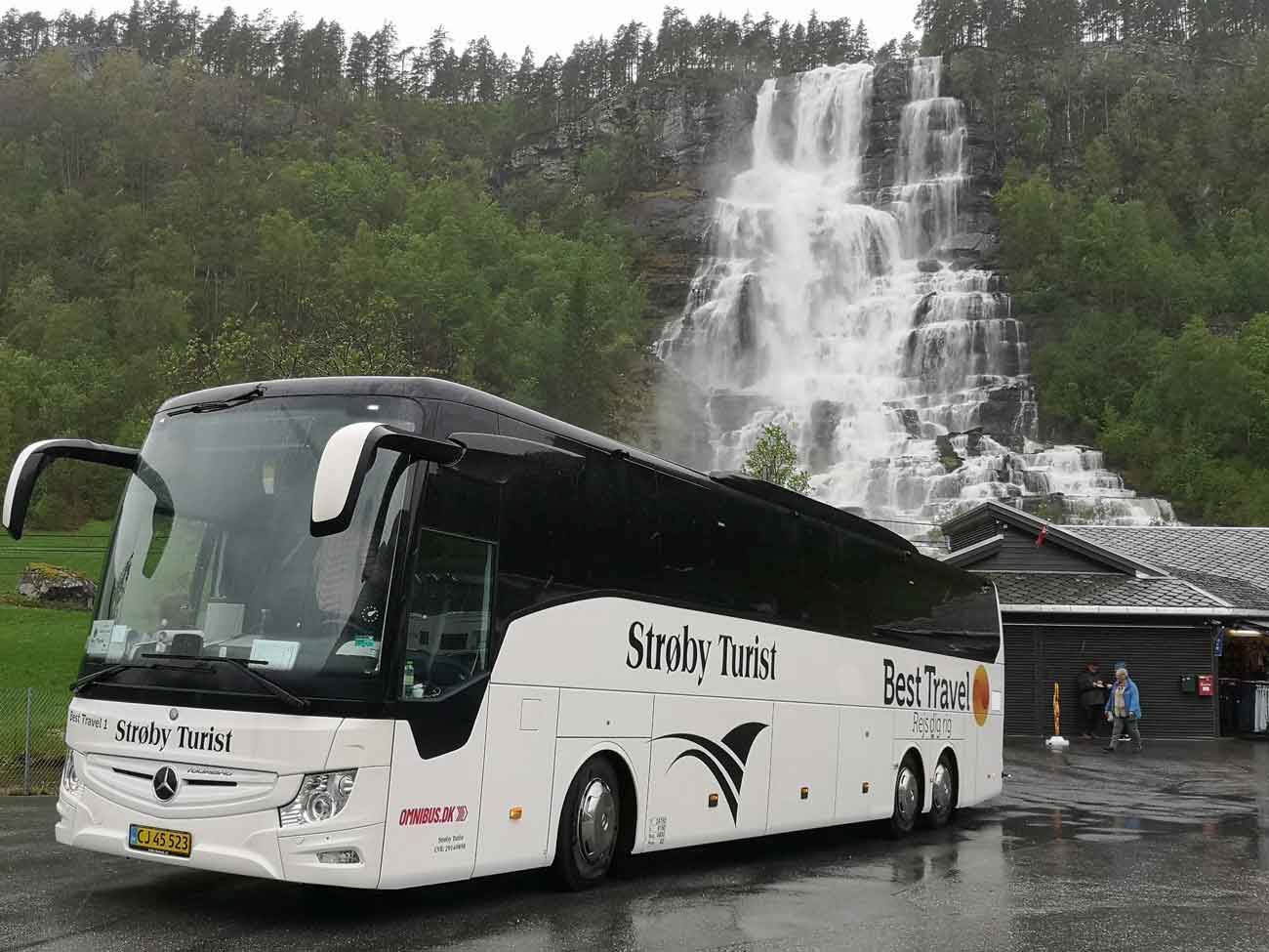 En hvid turistbus parkeret foran et vandfald i et frodigt, grønt landskab.