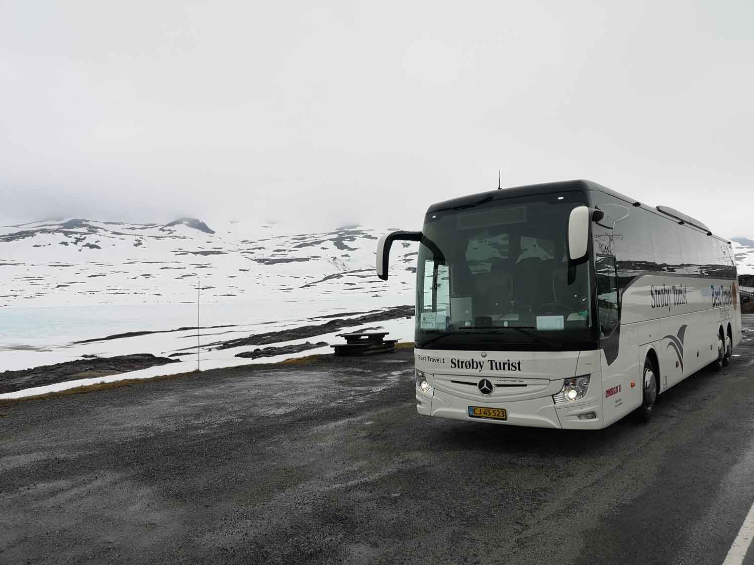 Hvid turistbus på en vej med snedækkede bjerge i baggrunden.