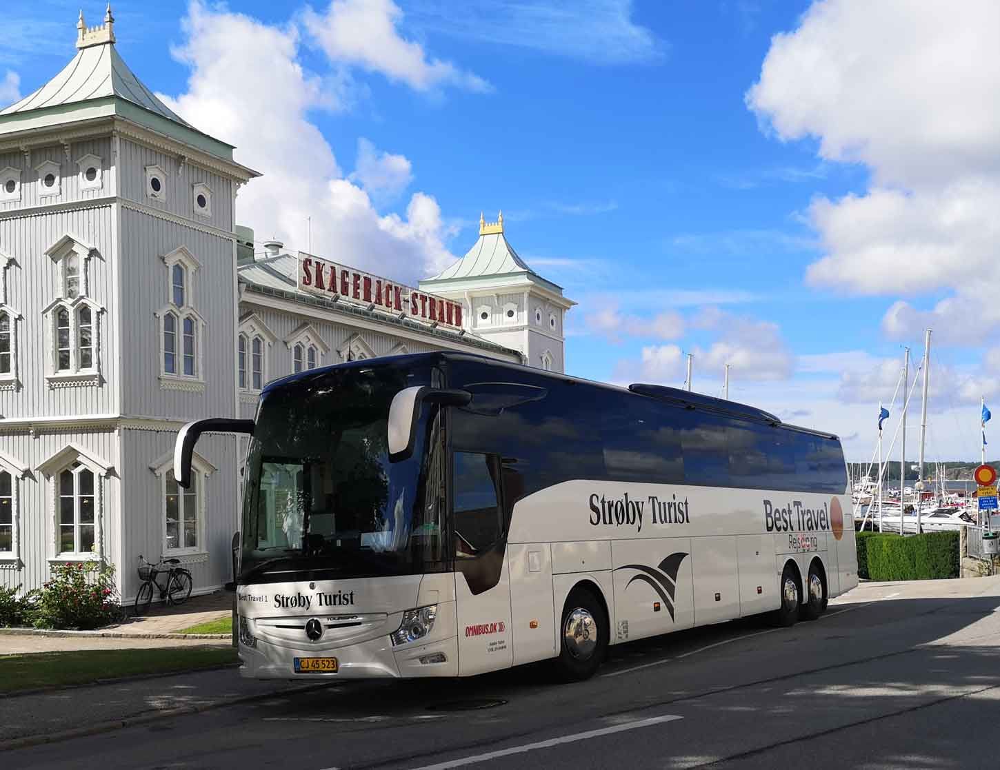 Hvid bus parkeret foran en lys bygning med grønt tag. Blå himmel, solskinsdag.