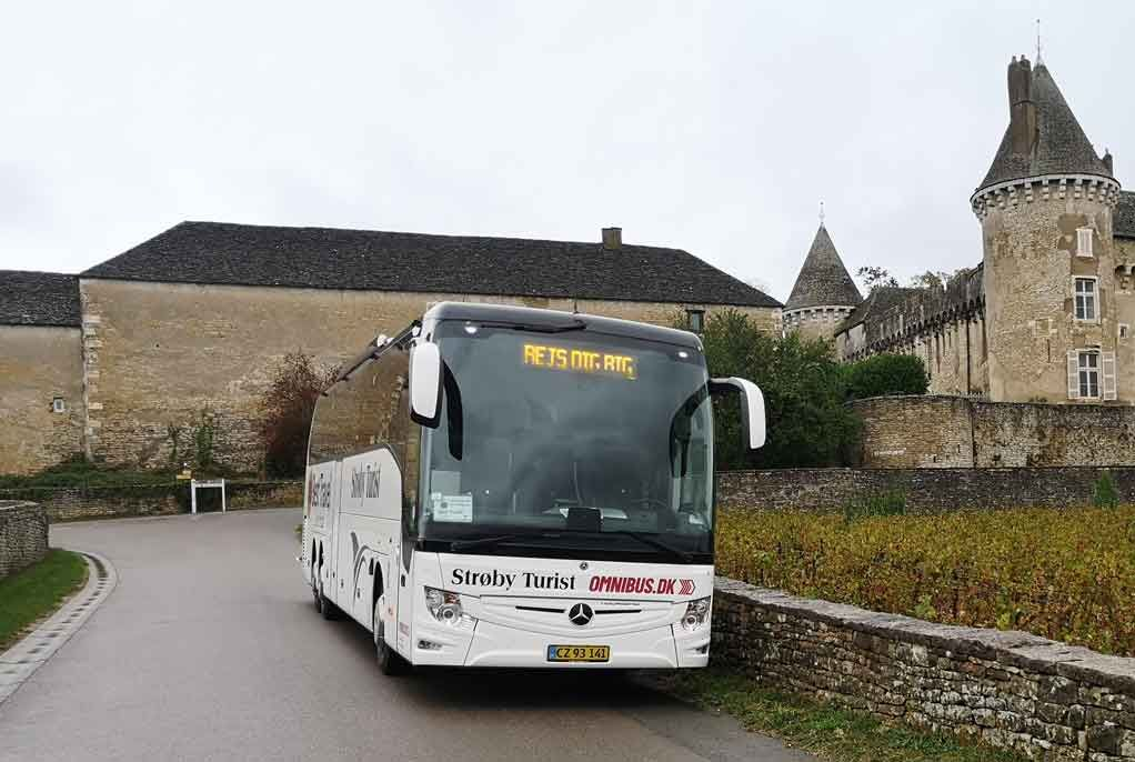 Hvid turistbus på en vej, parkeret foran et stenslot på en overskyet dag.
