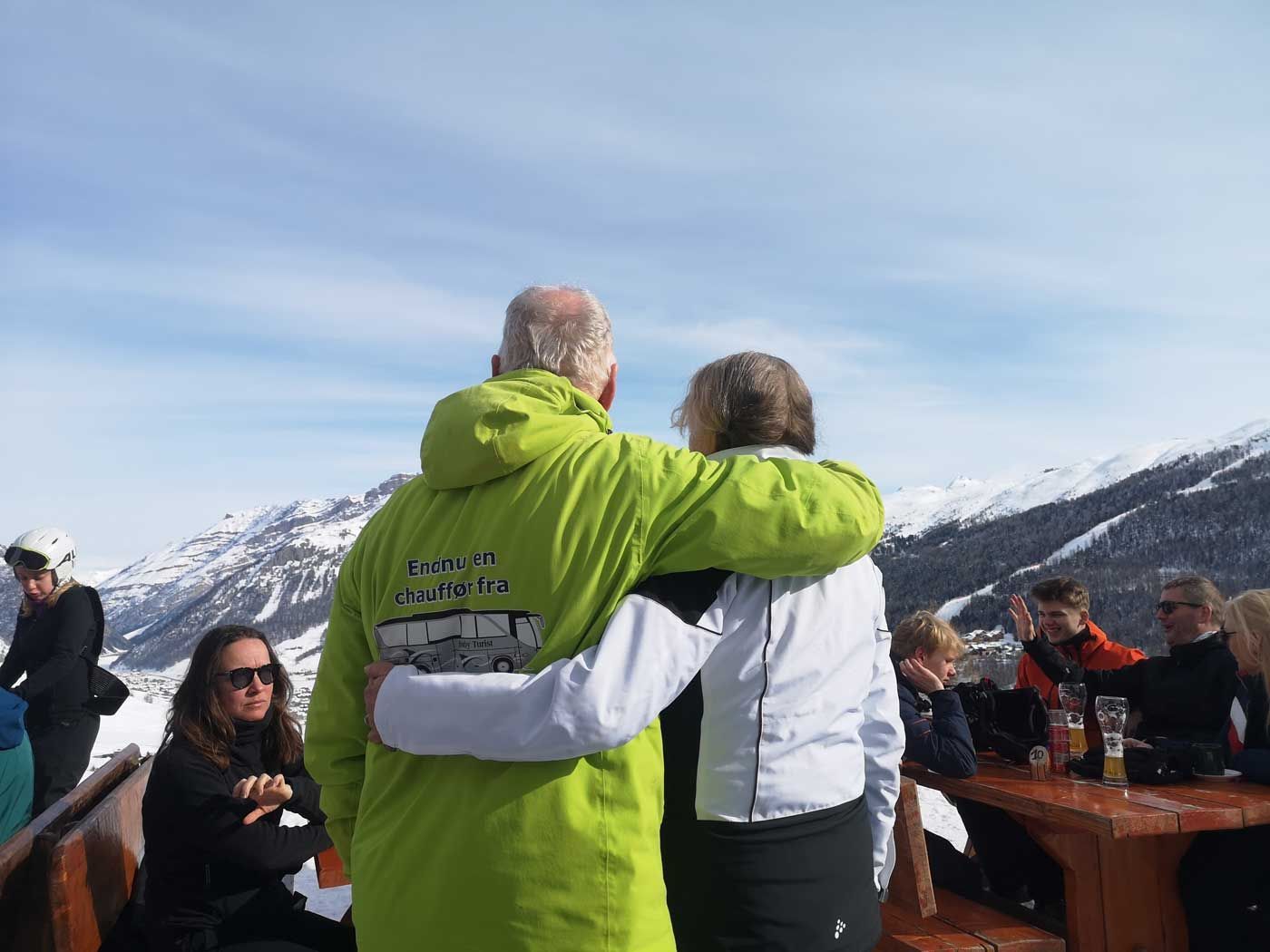 To personer med arme om hinanden, der kigger på sneklædte bjerge. Andre sidder ved et bord.