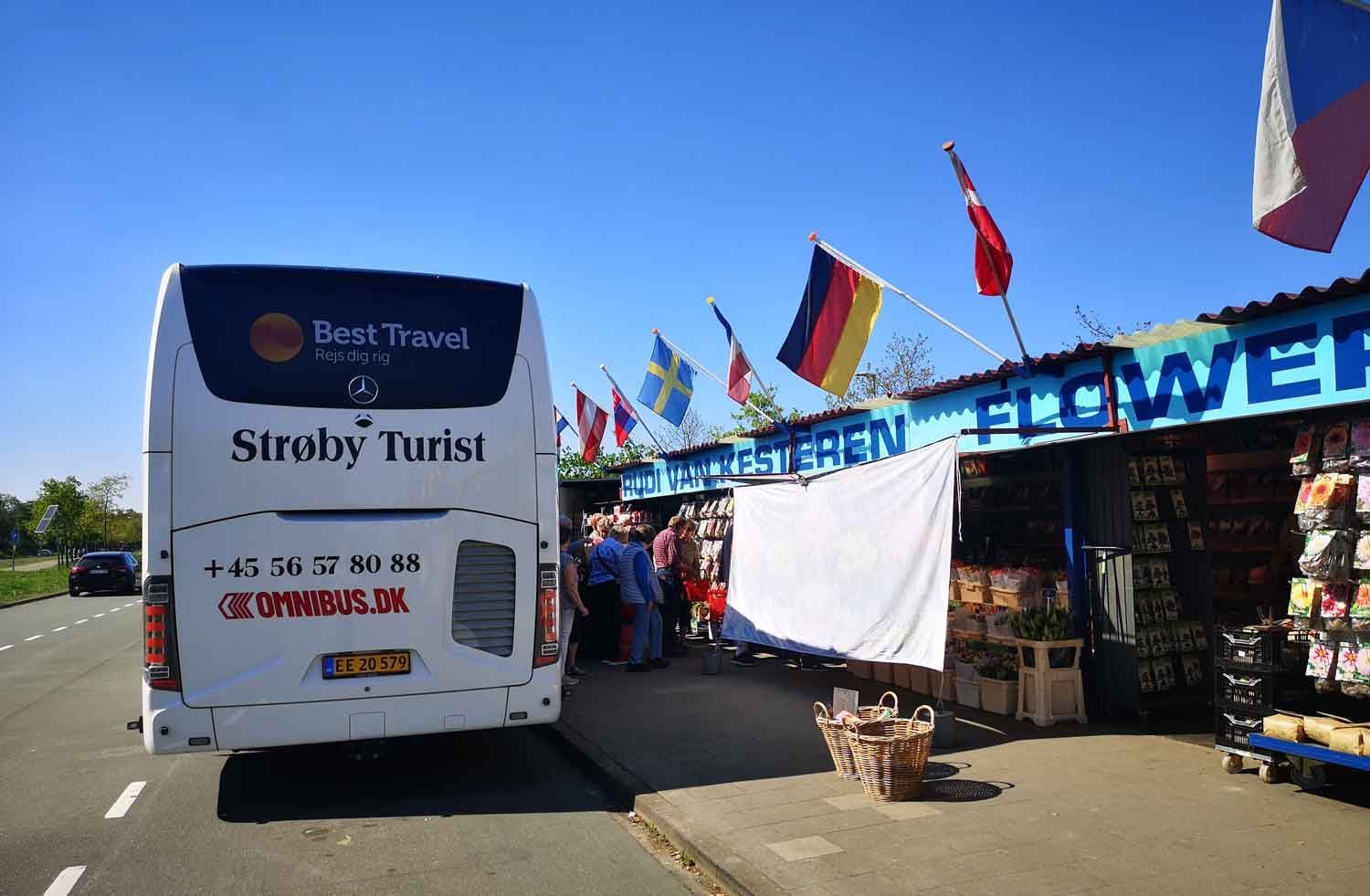 Bus parkeret nær en blomsterbod med flag. Folk samles ved boden under en blå himmel.