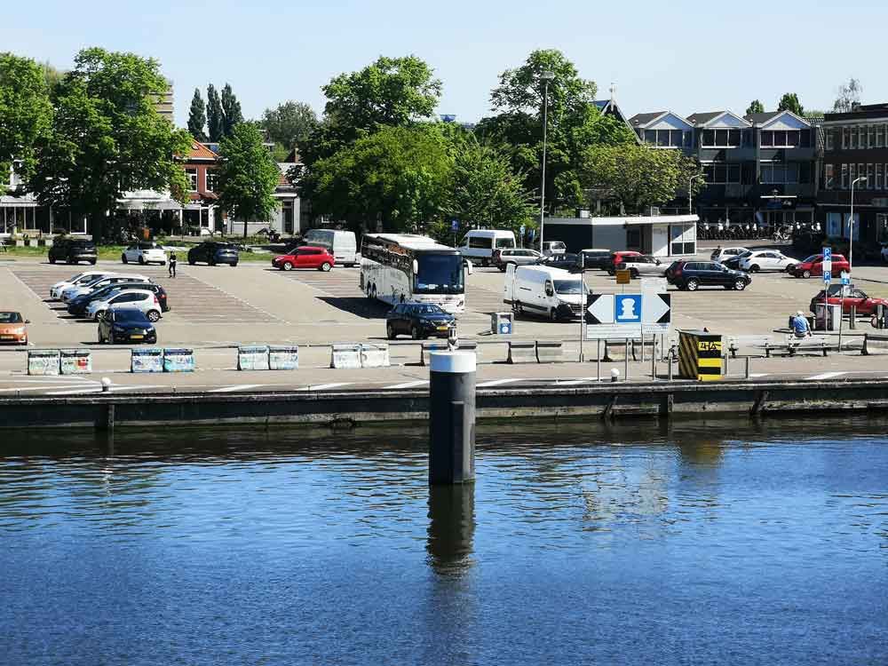 Parkeringsplads ved vandet med biler, busser og bygninger under en blå himmel.