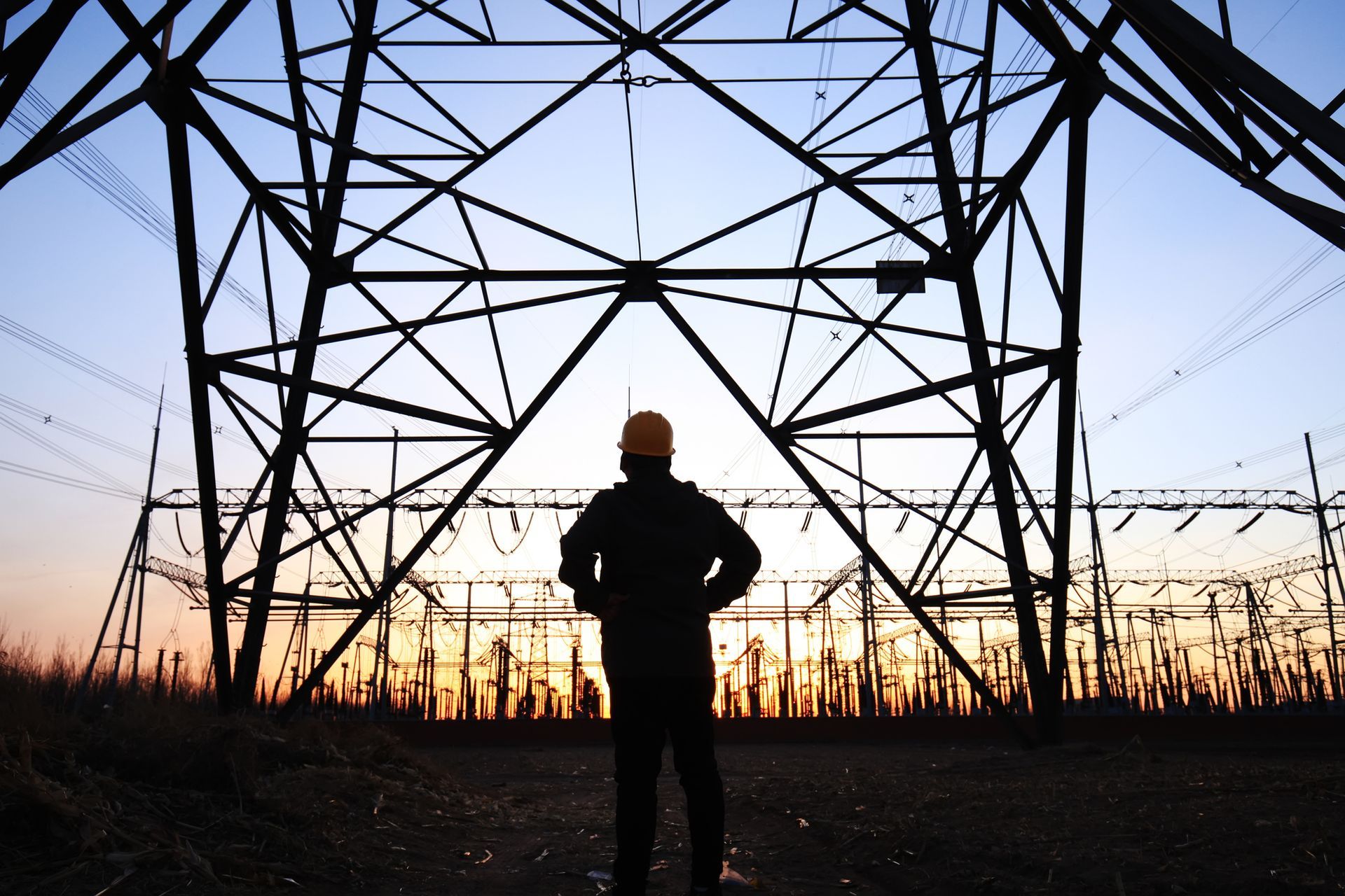 Silhouette of a worker in a hard hat, looking up at a tall electrical tower at sunset.