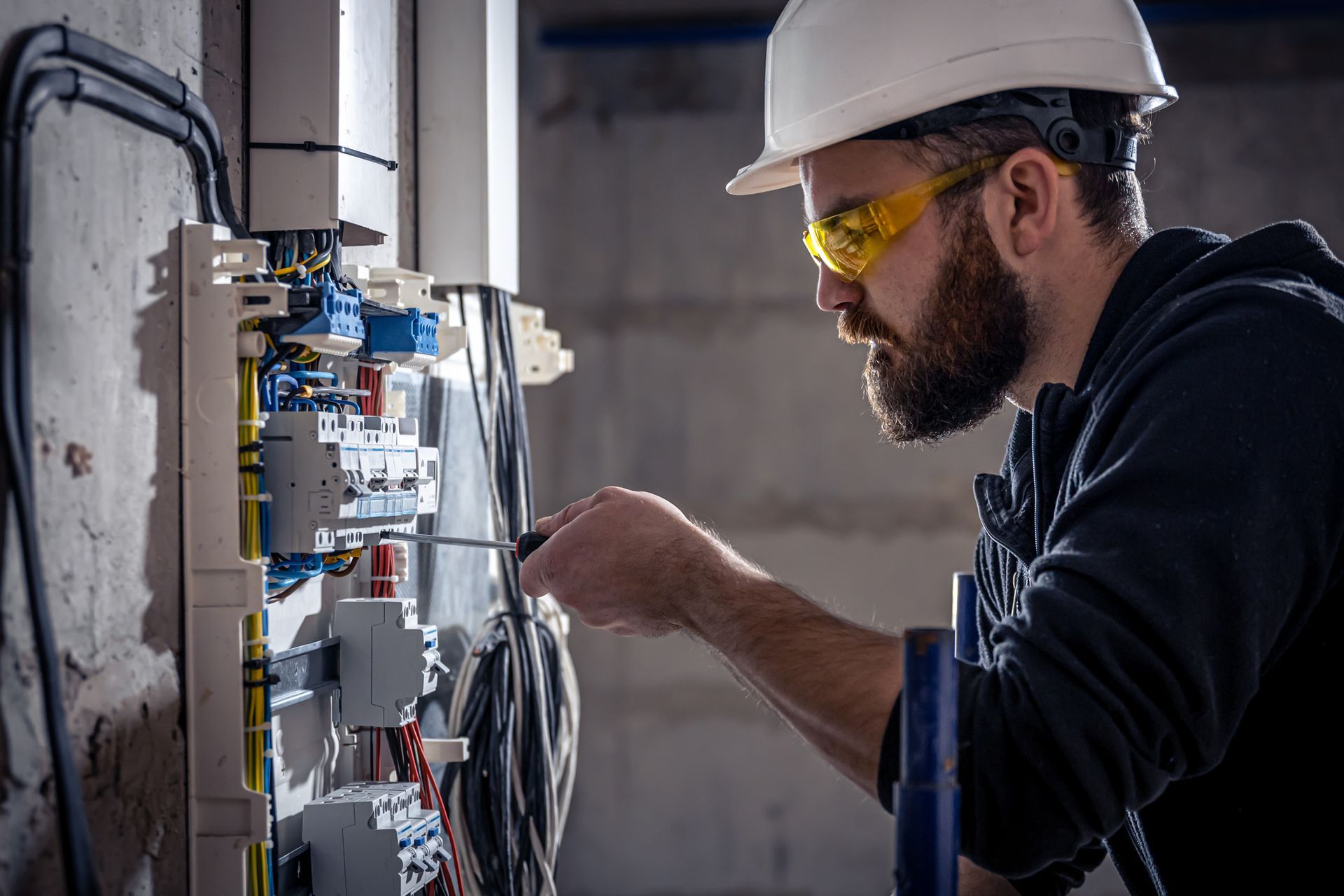 Electrician in hard hat and safety glasses, working on a circuit panel in a building.