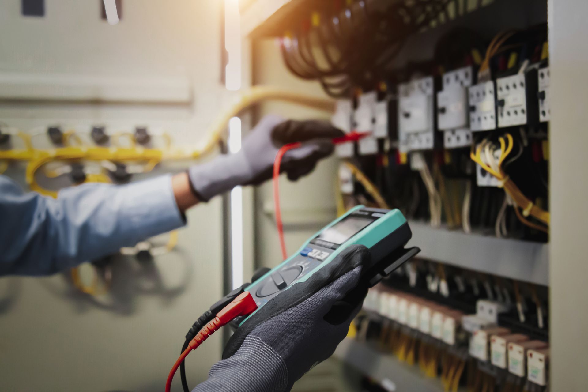 Electrician using a multimeter to check electrical panel; wearing gloves, indoor setting.