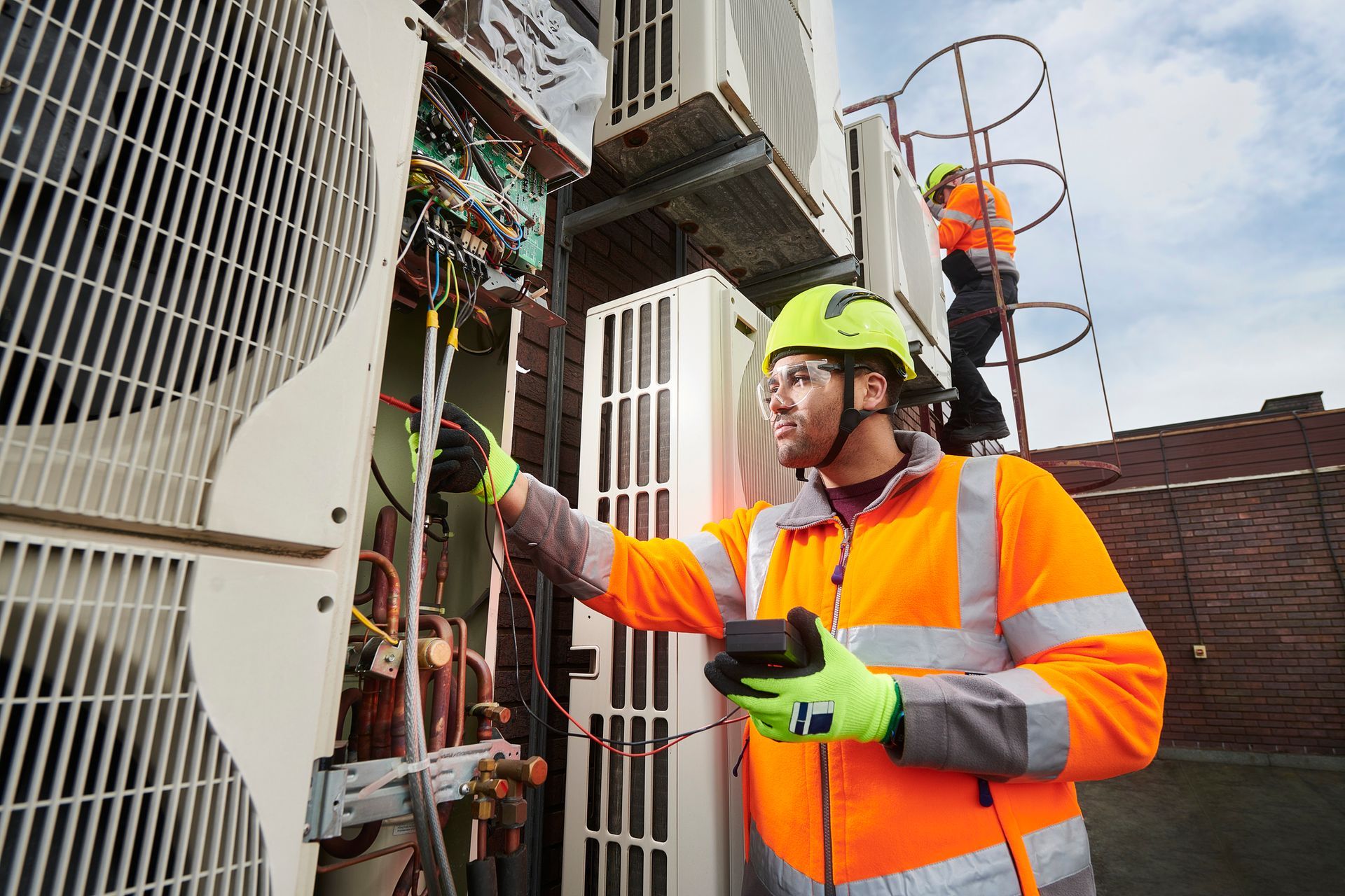 Two HVAC technicians in orange vests, one inspecting wiring, the other on a ladder, against a backdrop of rooftop air conditioning units. Two HVAC technicians in orange vests, one inspecting wiring, the other on a ladder, against a backdrop of rooftop air conditioning units.