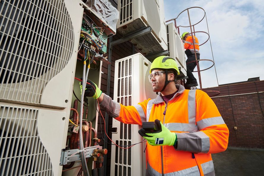 Two HVAC technicians in orange vests, one inspecting wiring, the other on a ladder, against a backdrop of rooftop air conditioning units.