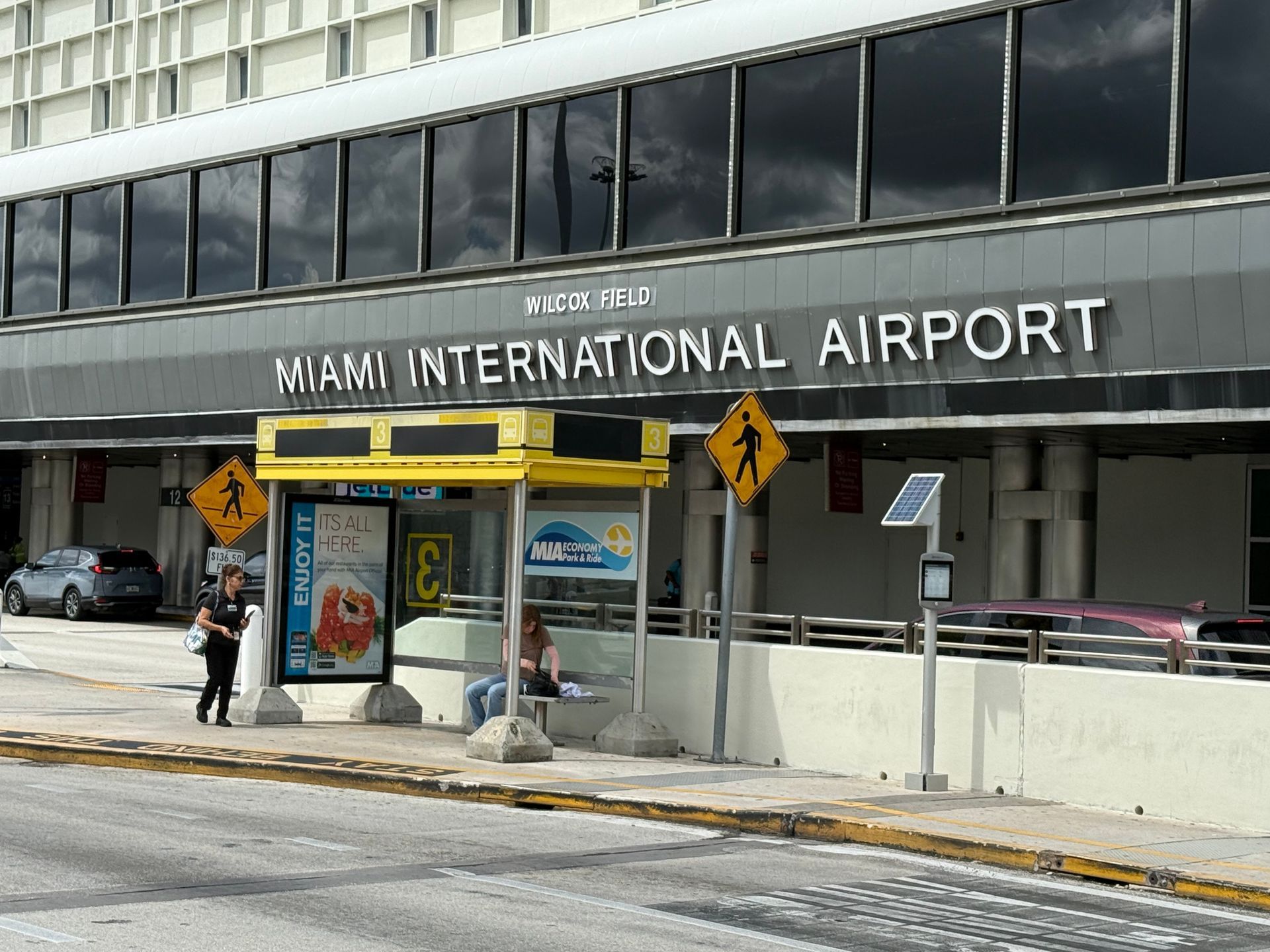 Miami International Airport exterior with bus stop, people, and a car.