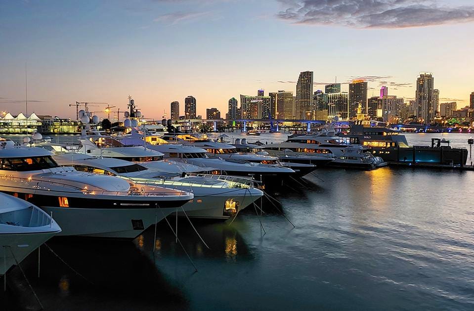 A row of boats are docked in a harbor with a city skyline in the background.