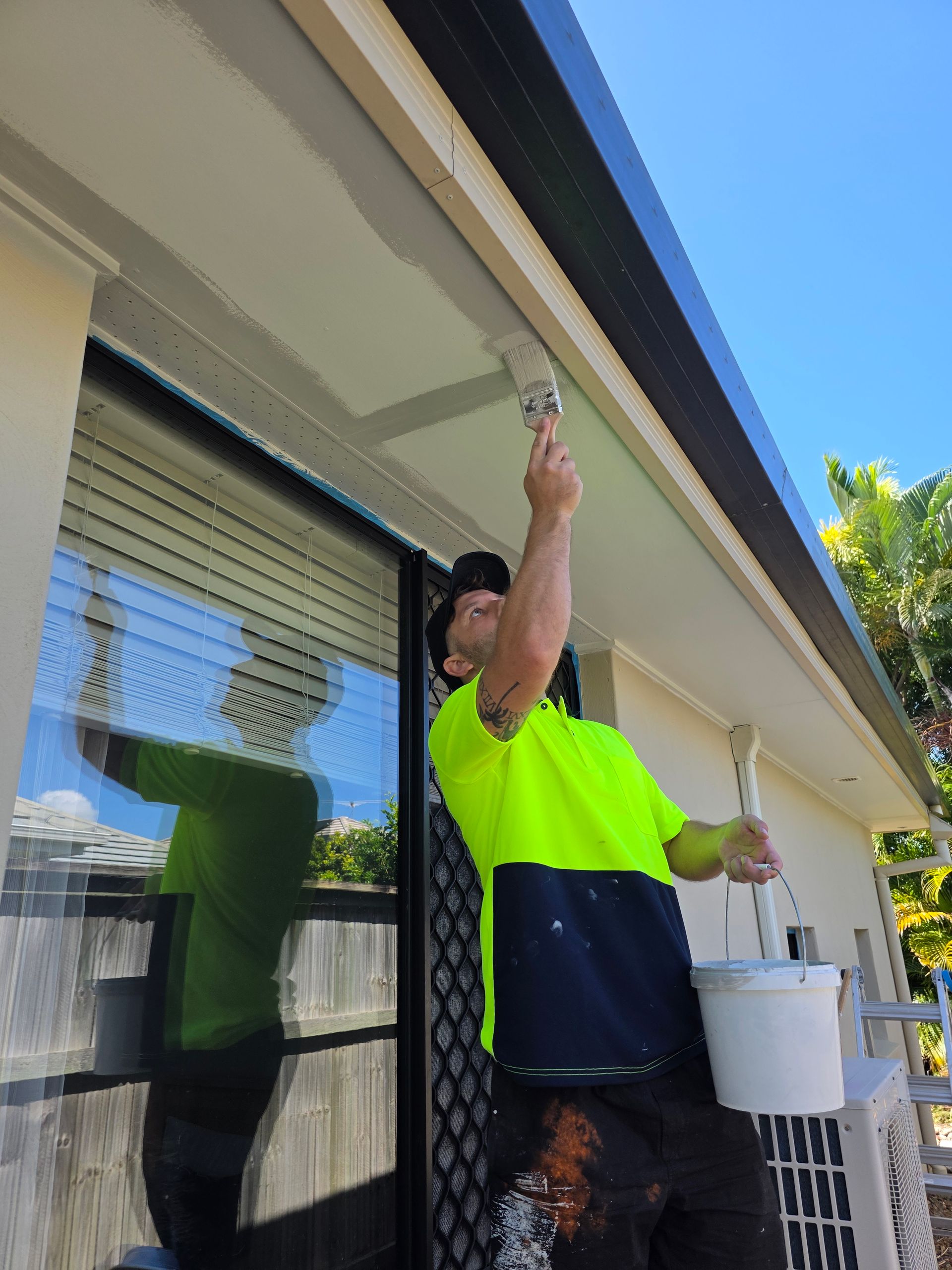 Man painting the exterior of a building's trim, standing on a ladder. Wearing work clothes, a white bucket nearby.