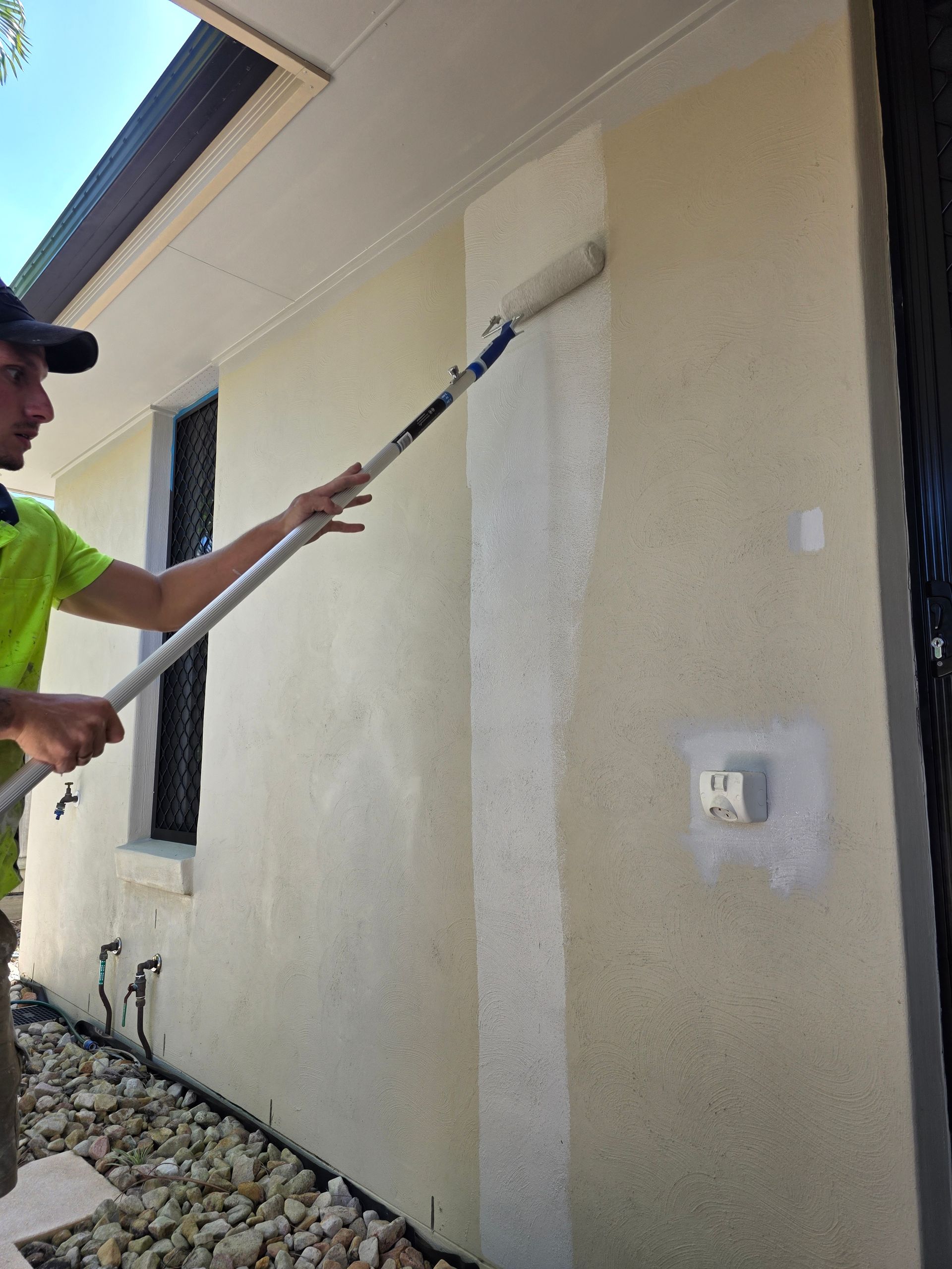 Man painting exterior wall with a roller. Wall is beige; man wears a hat and green shirt.