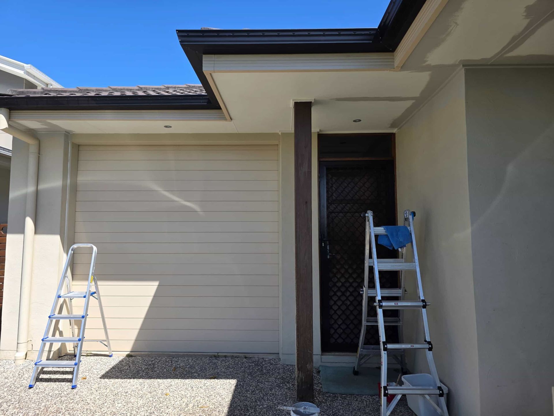 Exterior of house with garage and door, two ladders. Beige garage door and walls, brown gutters and pillar. Blue sky. — Legacy Surface Solutions in Mango Hill, QLD