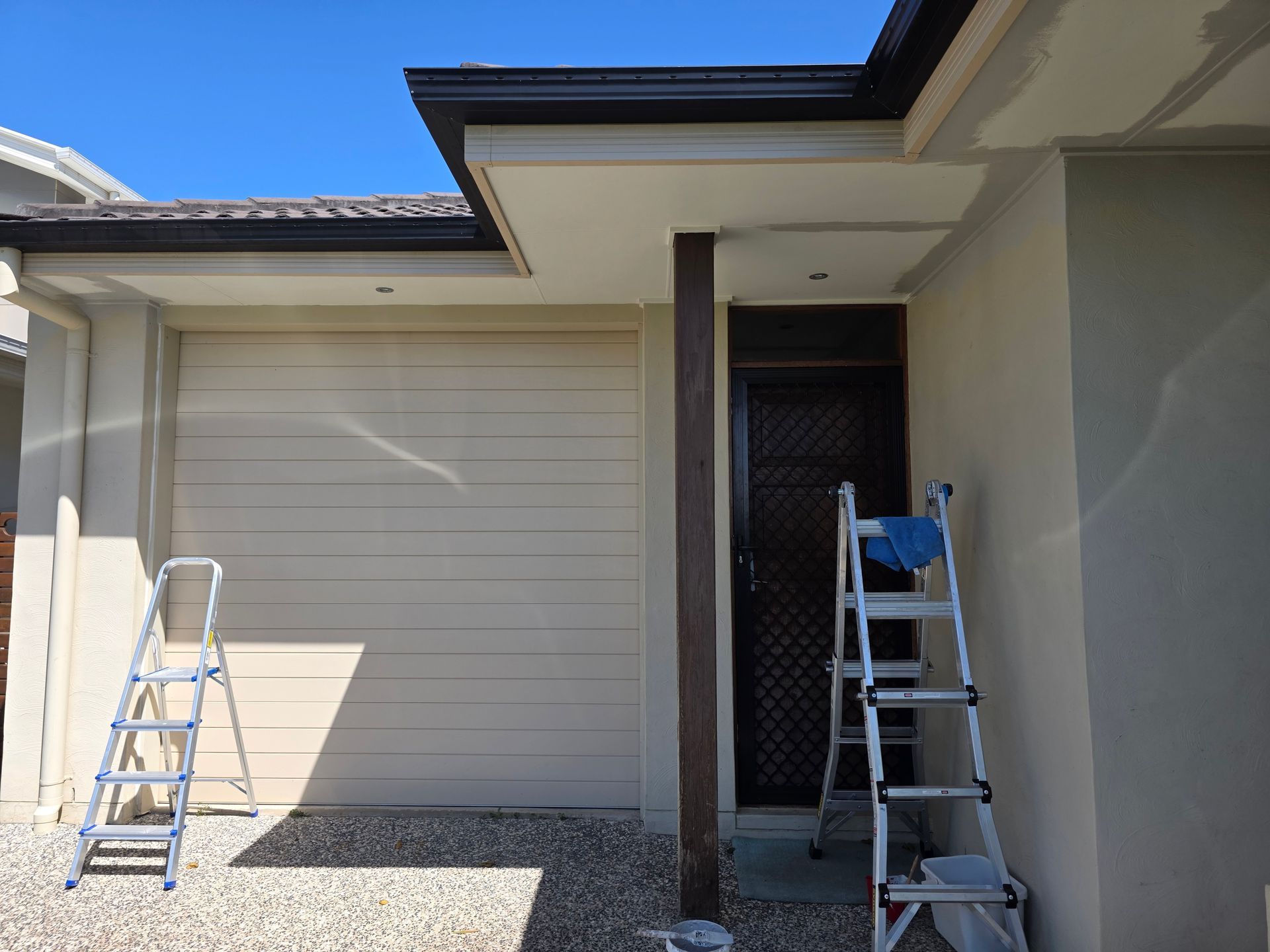 Exterior view of a house with a garage and front door, two ladders and gravel ground.