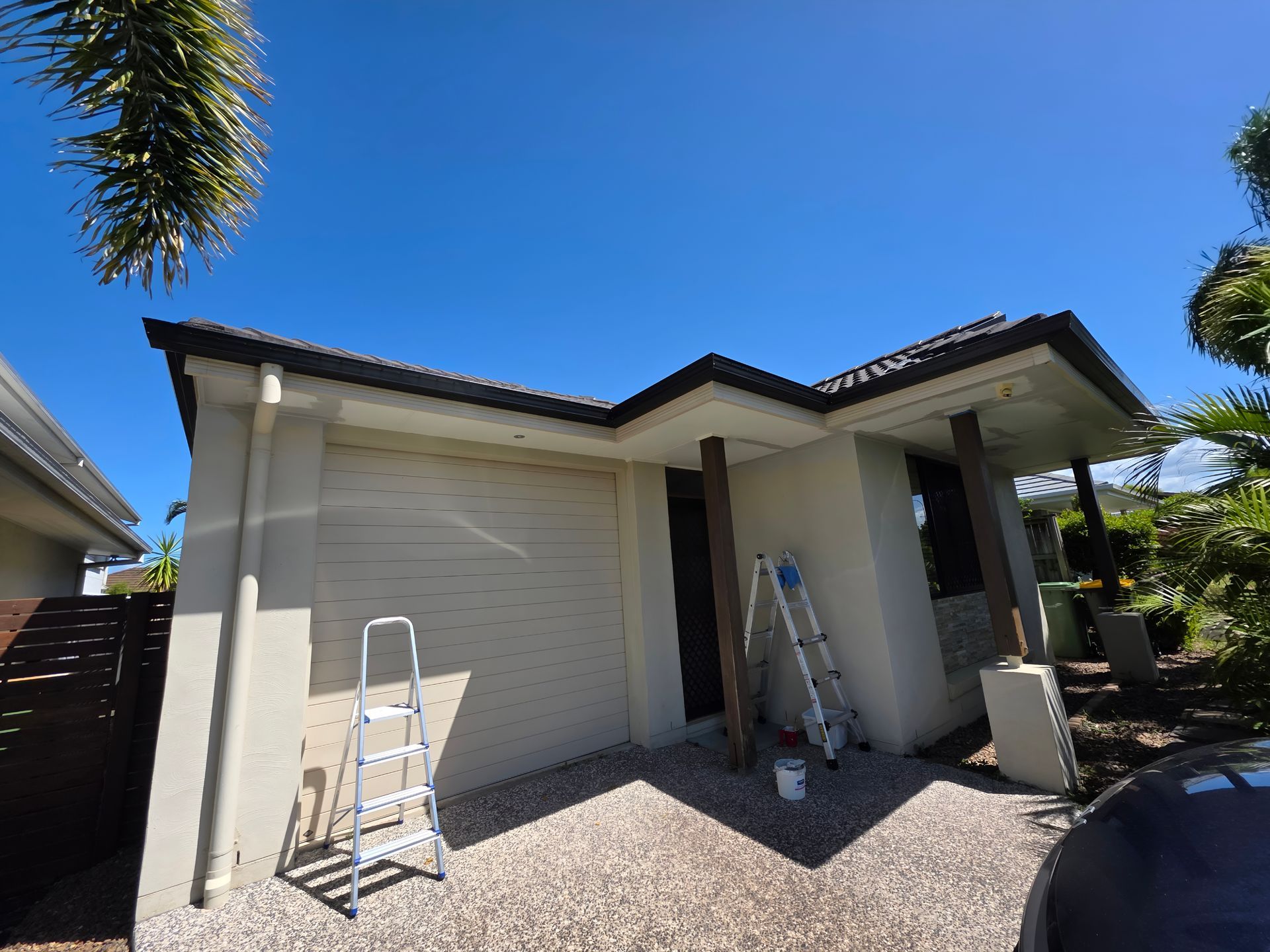 Beige house exterior with dark roof. Ladders, gravel driveway, and clear blue sky.