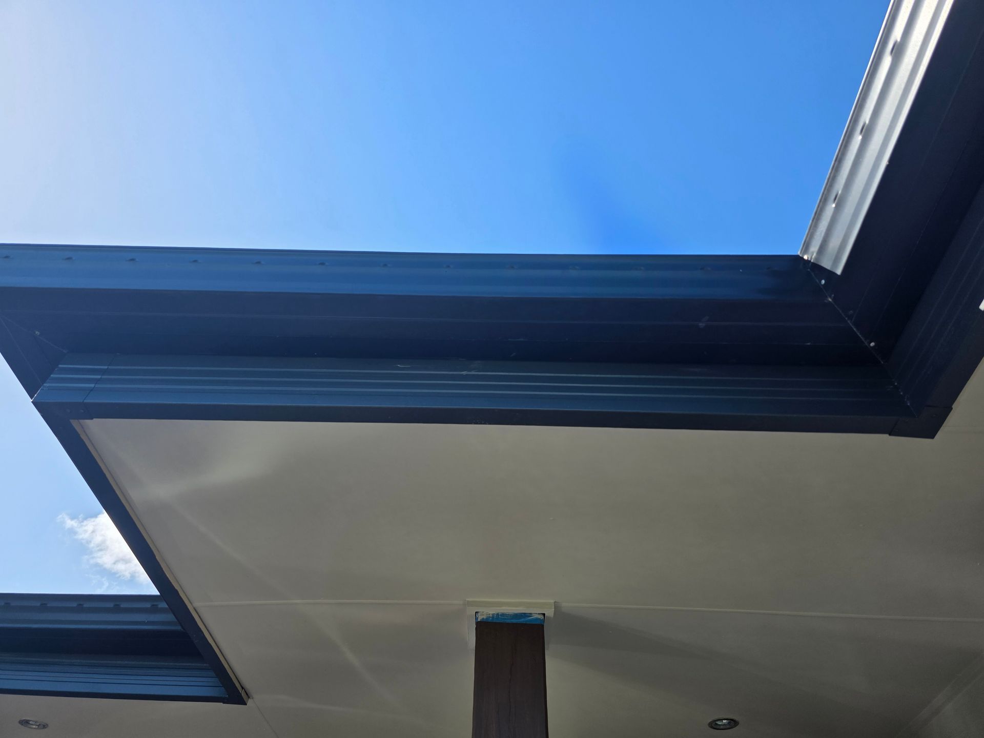 Low-angle view of a roof's edge against a blue sky; dark fascia and gutters, white soffit, and wooden support.