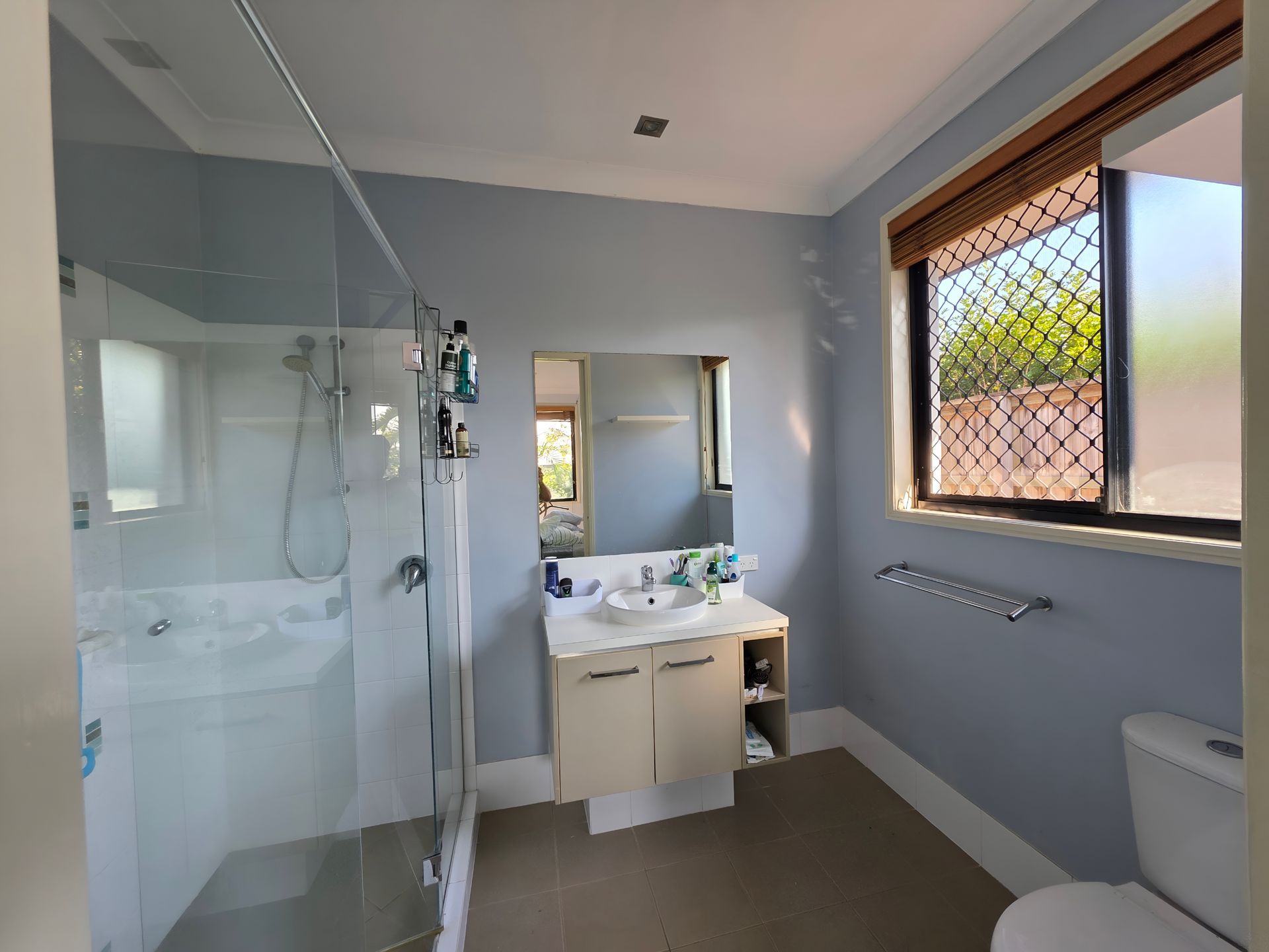 Bathroom with a shower, sink, toilet, and window with a view. Light blue walls, tan floor.