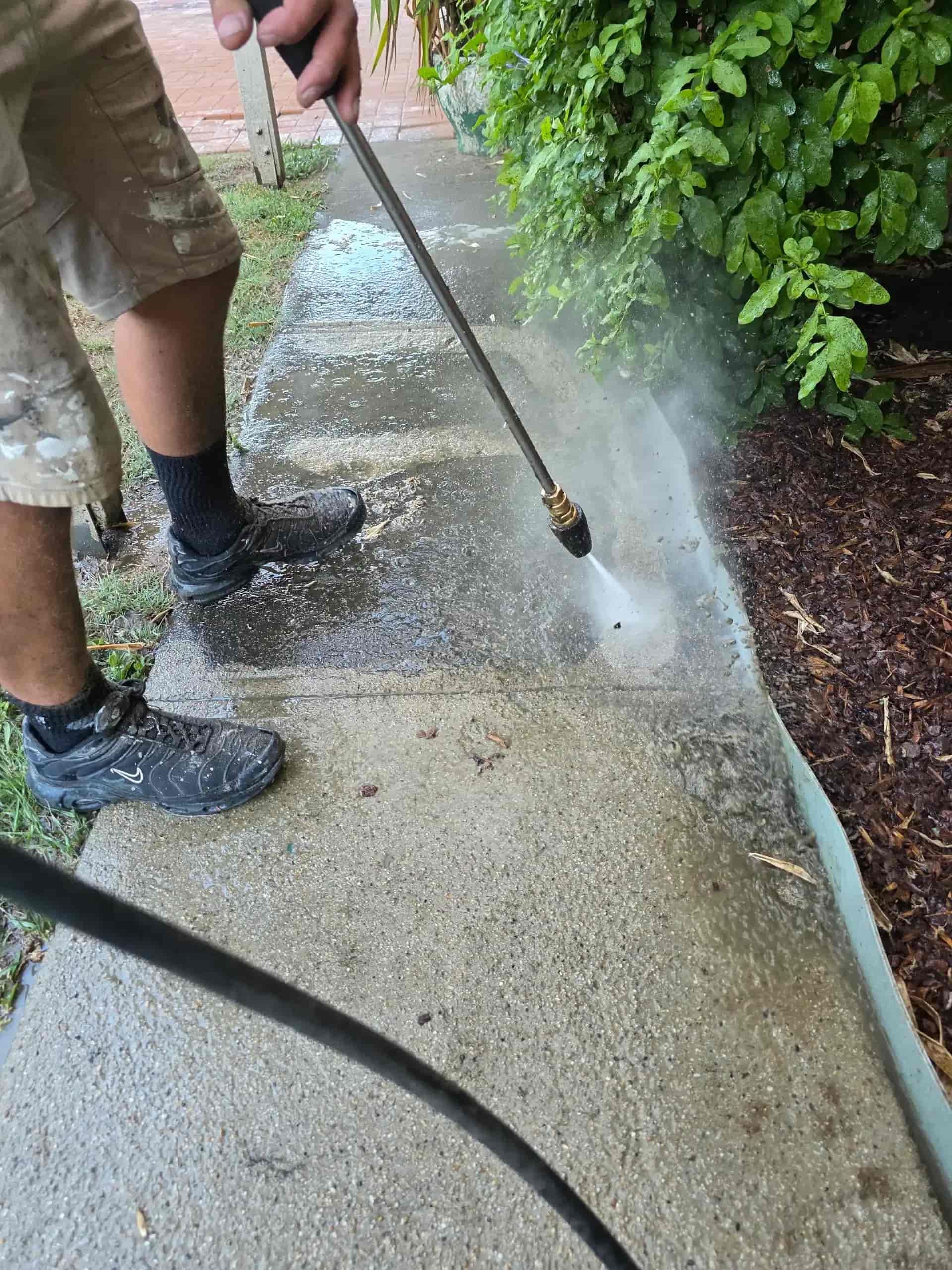 Person pressure washing a concrete sidewalk next to a bed of mulch and greenery. — Legacy Surface Solutions in Mango Hill, QLD