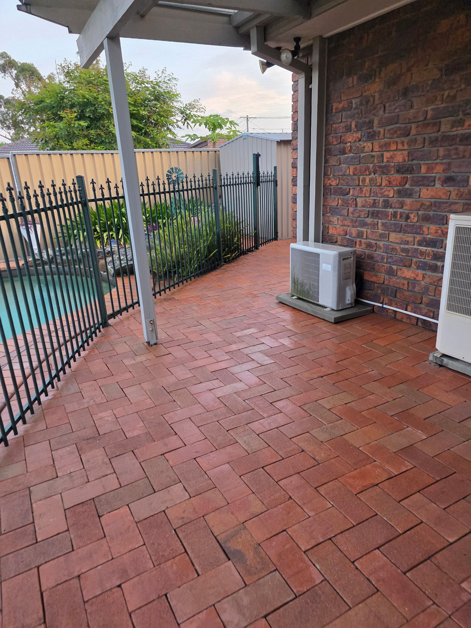 Brick patio next to pool and house with black fence. Air conditioners on the wall.