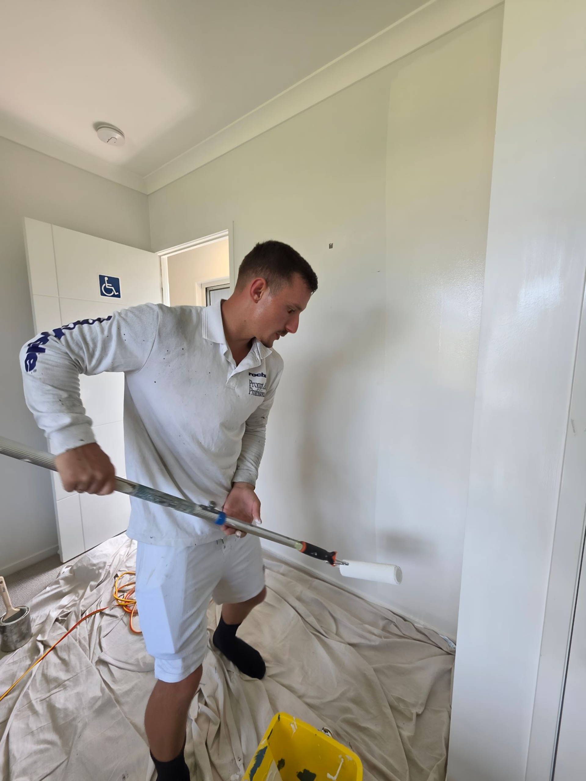 Man painting a white wall with a roller. Indoors, paint bucket visible, drop cloth on floor.