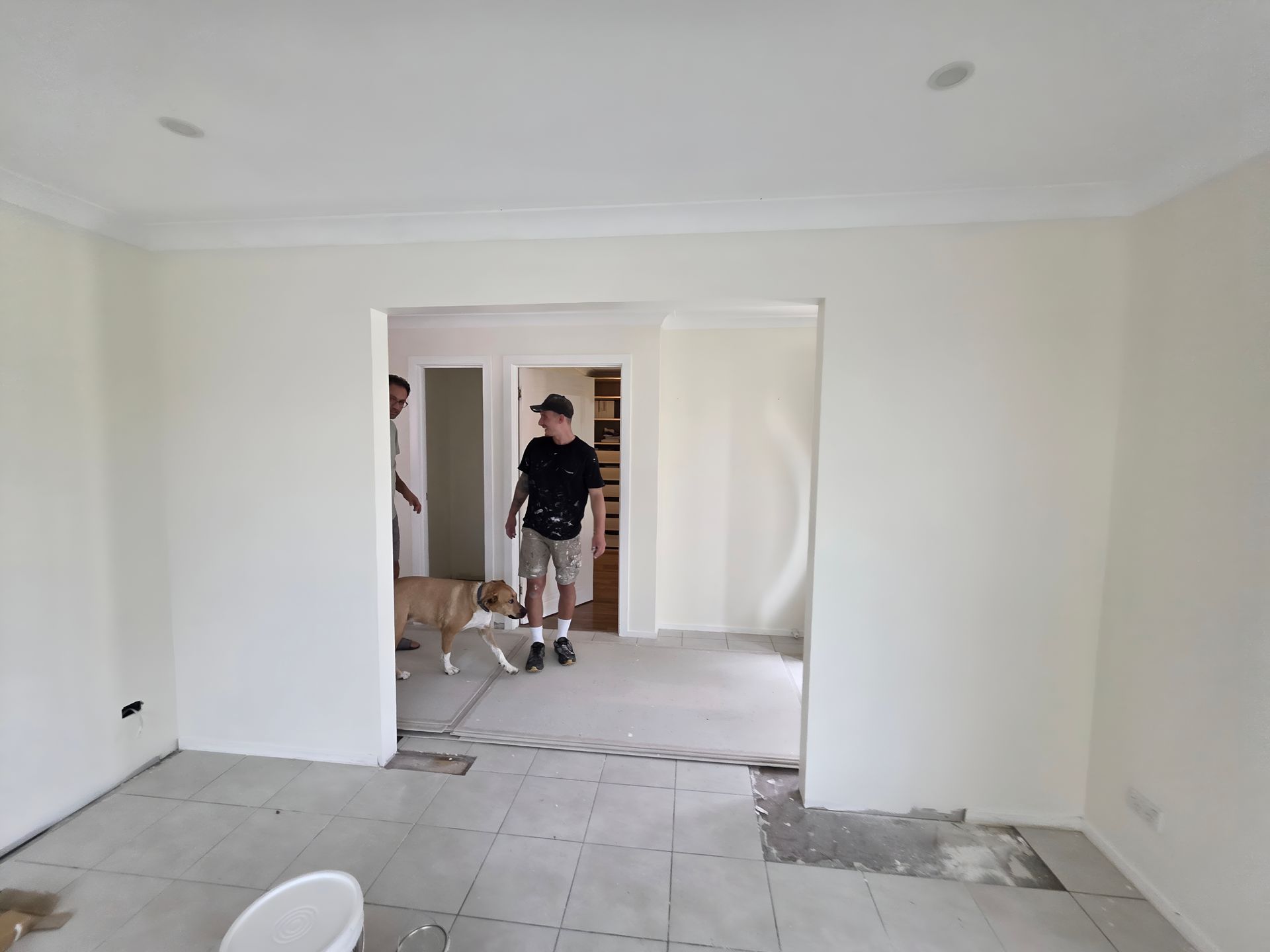 Room under renovation with two men, a dog, and missing floor tiles. White walls, doorway.