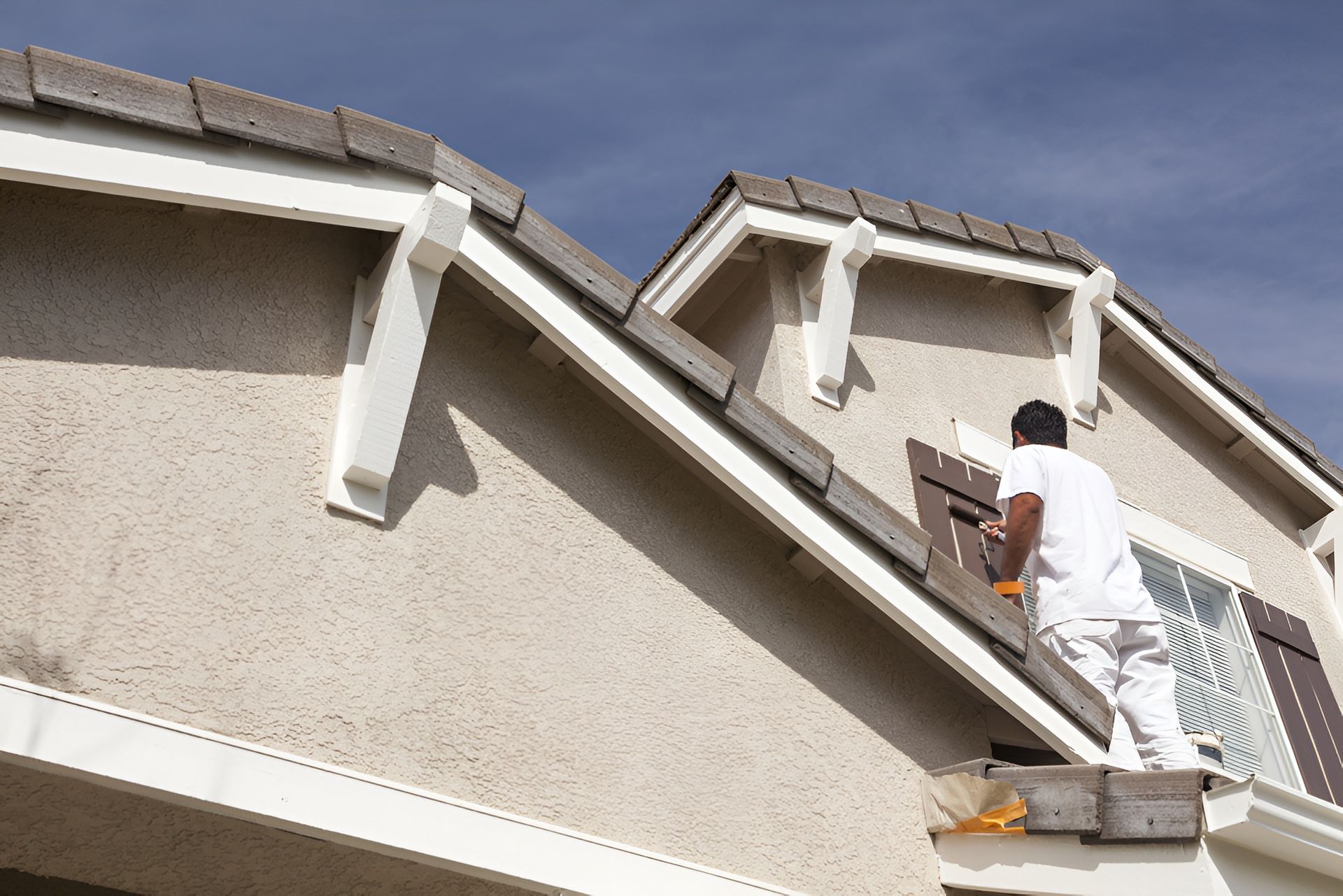 Exterior house painter applying paint to upper wall and roofline of a residential home