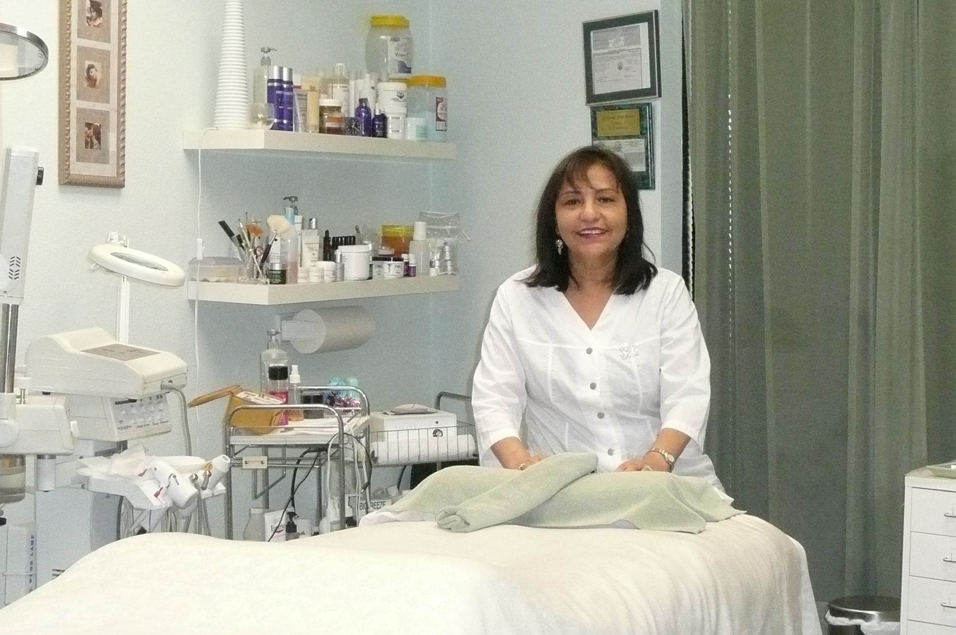 Woman in white coat standing near massage table in a spa-like room with equipment and shelves.