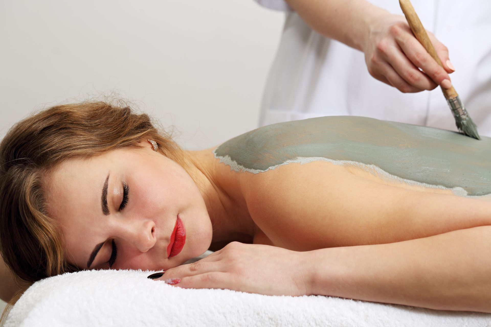 Woman receiving a mud back treatment at a spa; technician applying mud with a brush.