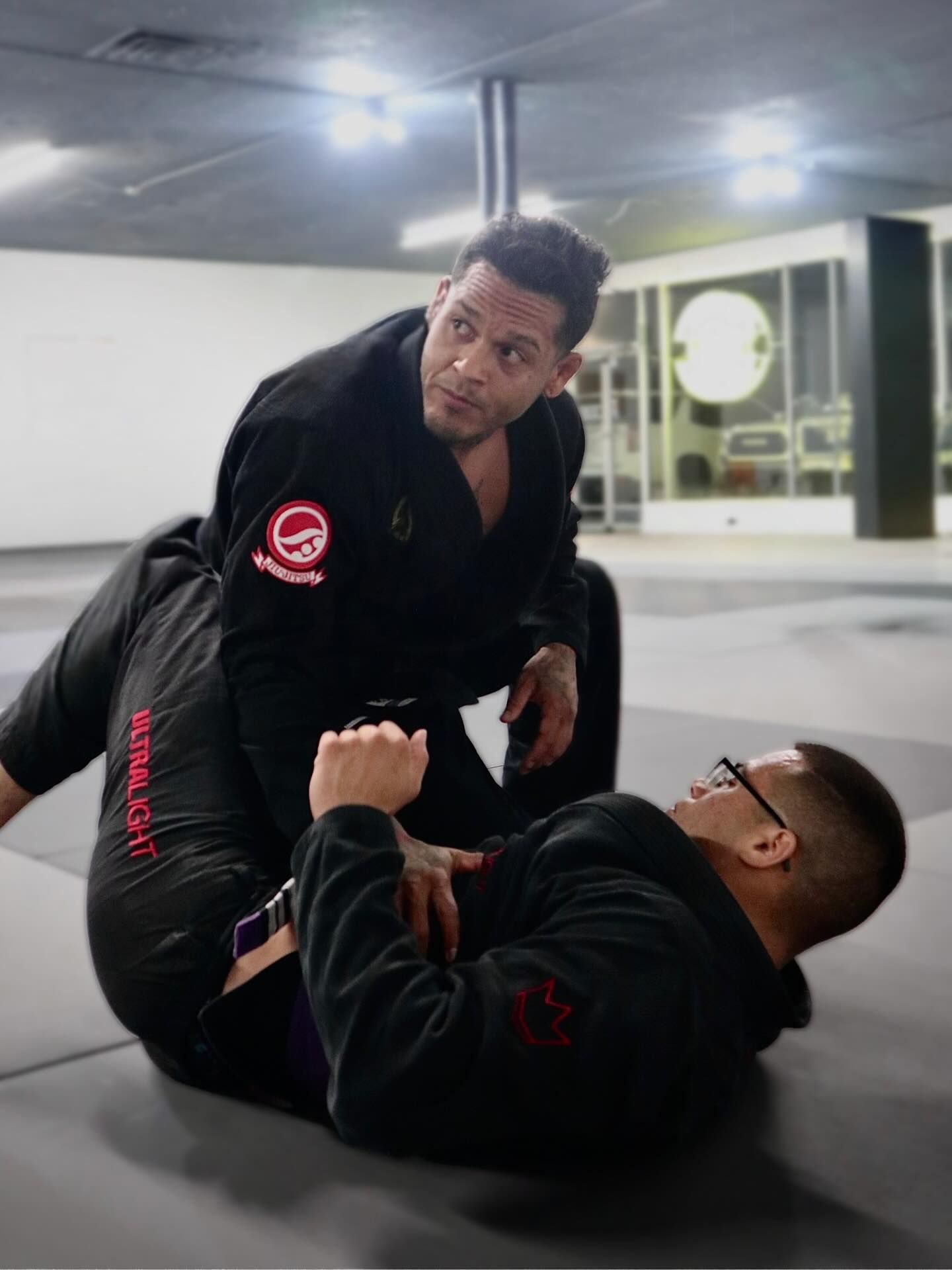 Two people grappling on a mat, one in a blue gi, the other in white. Gym setting.
