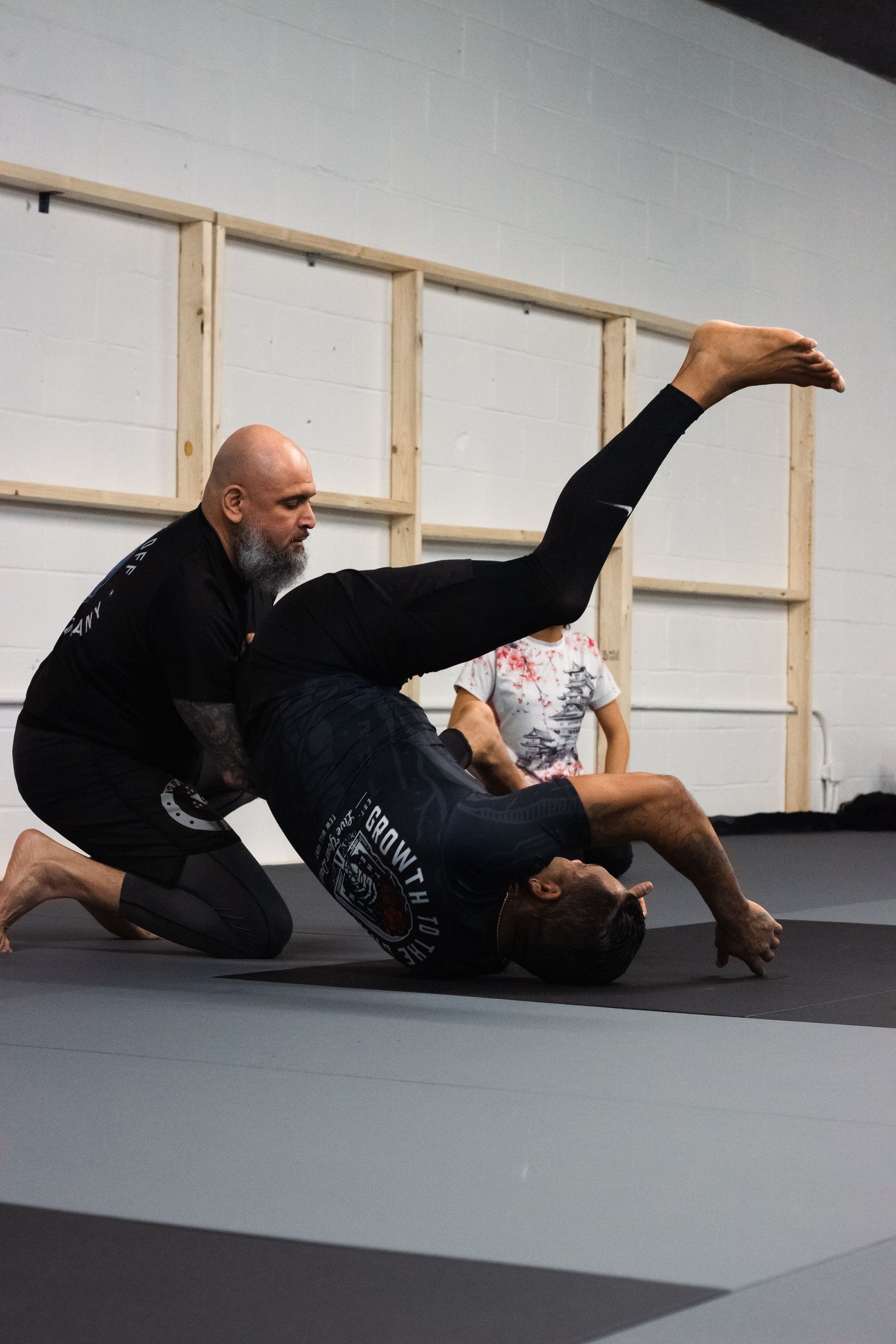 Two people in black and purple martial arts uniforms practice on blue mats in a gym. One is on top, applying pressure.