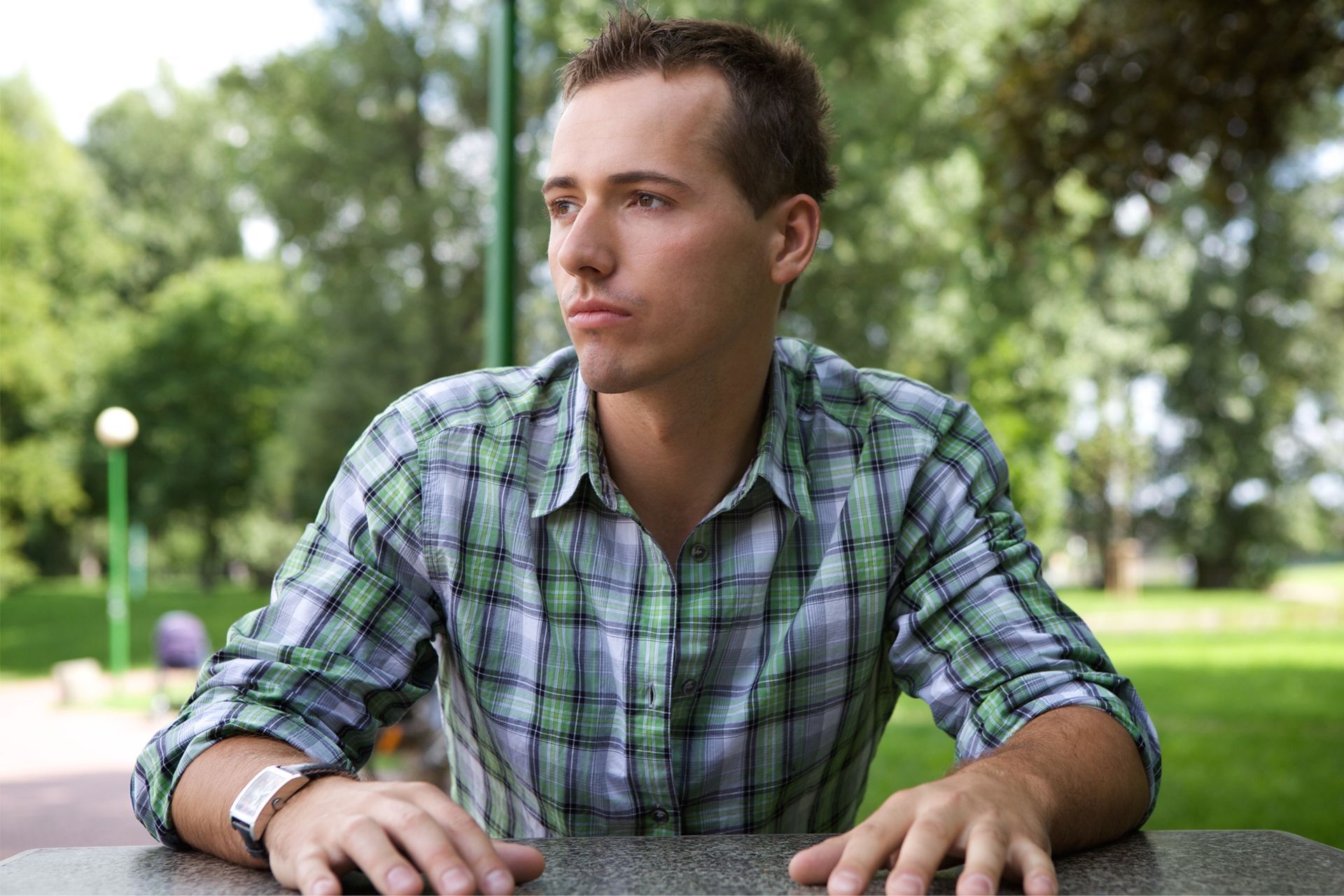 Man in plaid shirt sits at a table outdoors, looking off to the side with a serious expression.