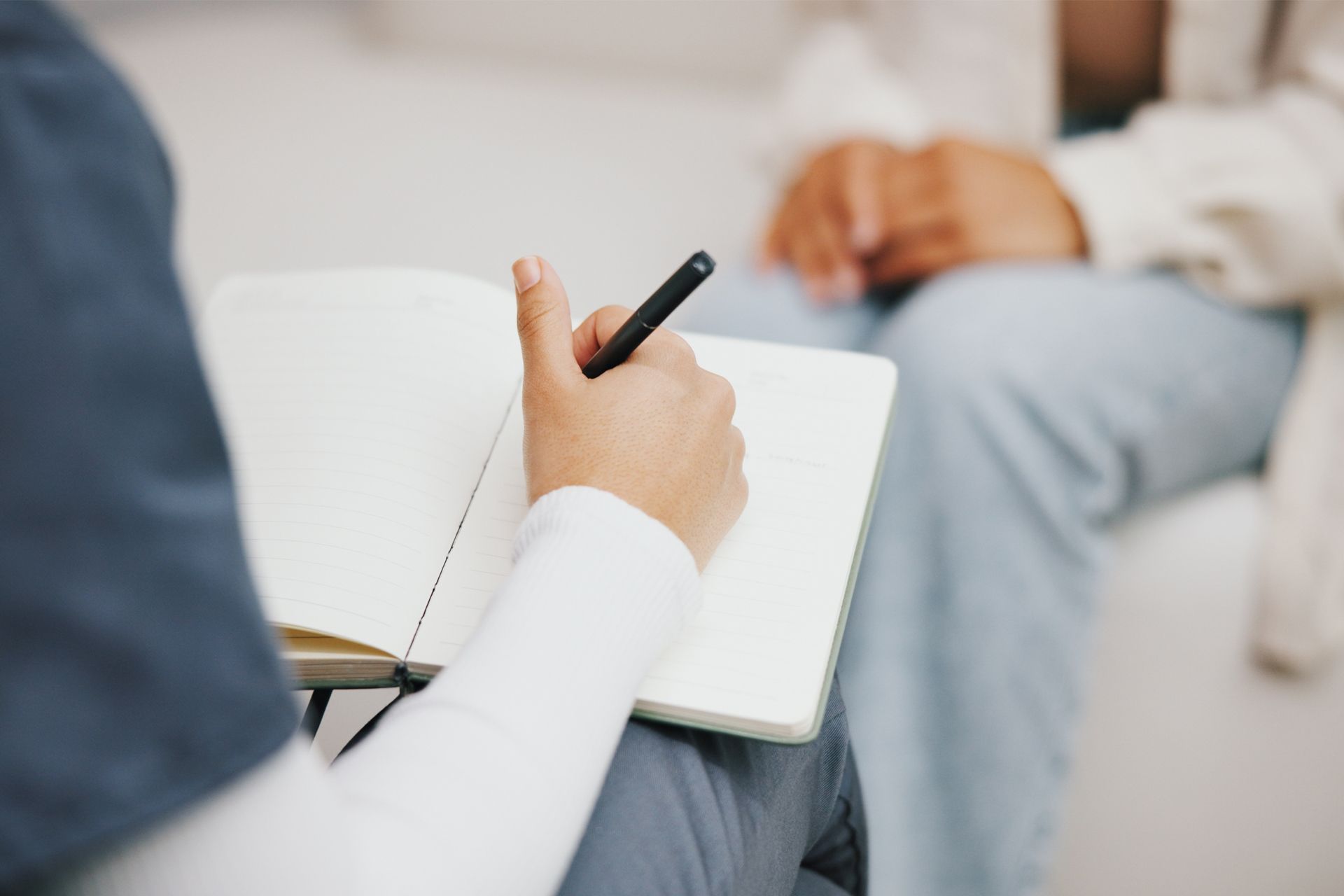 A person writing notes in a notebook while sitting across from someone in jeans.