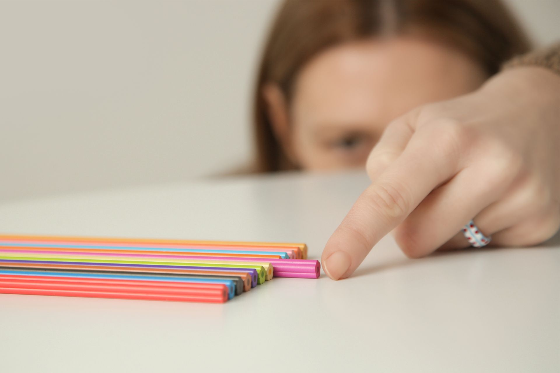 Person pointing at a pink pen on a white table next to a row of colorful pens.
