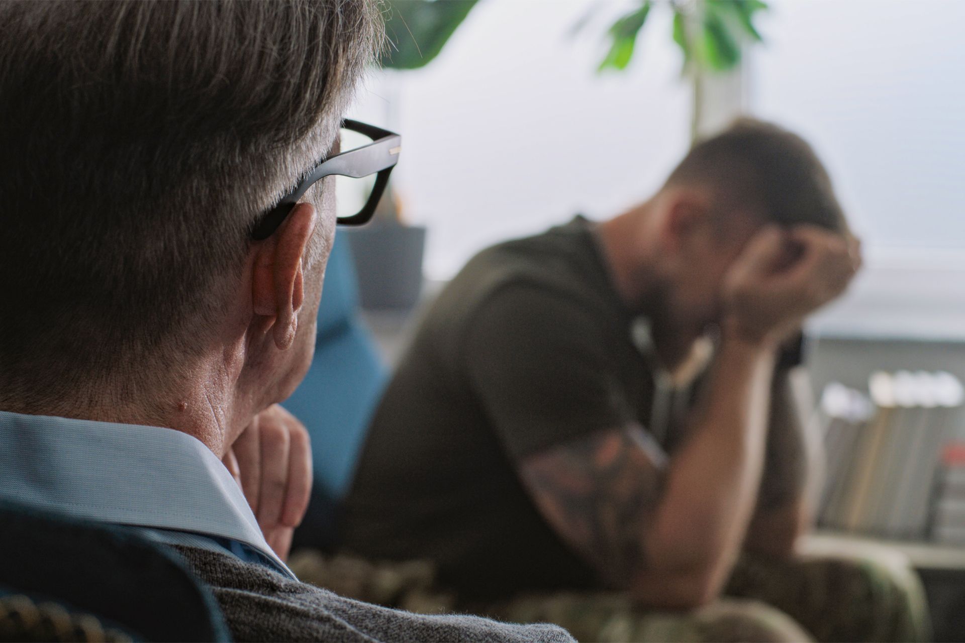 Therapist in glasses listens to a distressed person with head in hands; indoor setting.