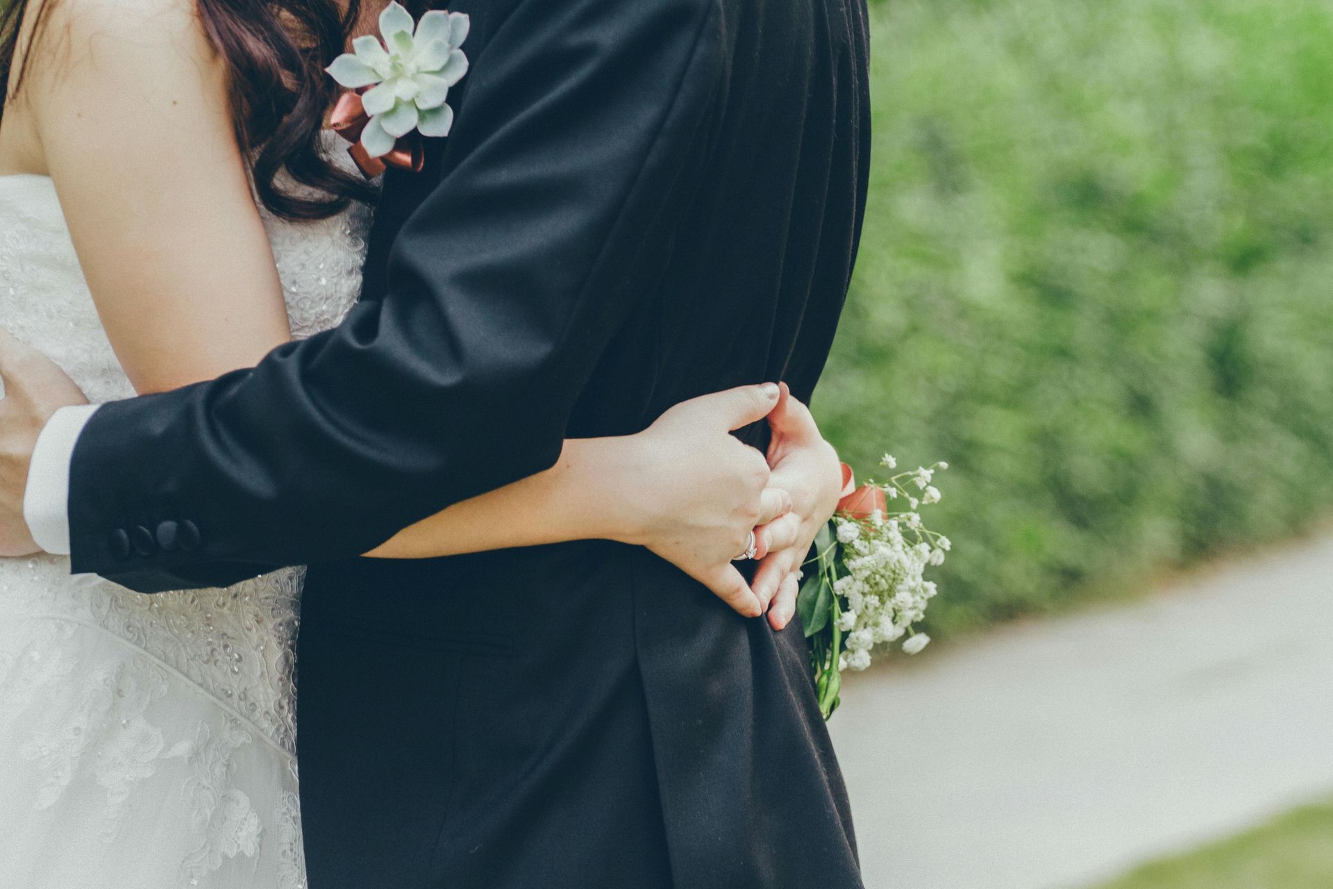 Bride and groom embrace outdoors, she in a white dress, he in a black suit.
