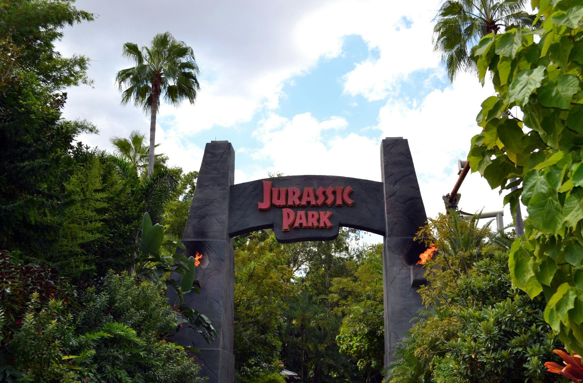 Jurassic Park entrance archway with flaming torches, surrounded by lush green foliage under a cloudy sky.