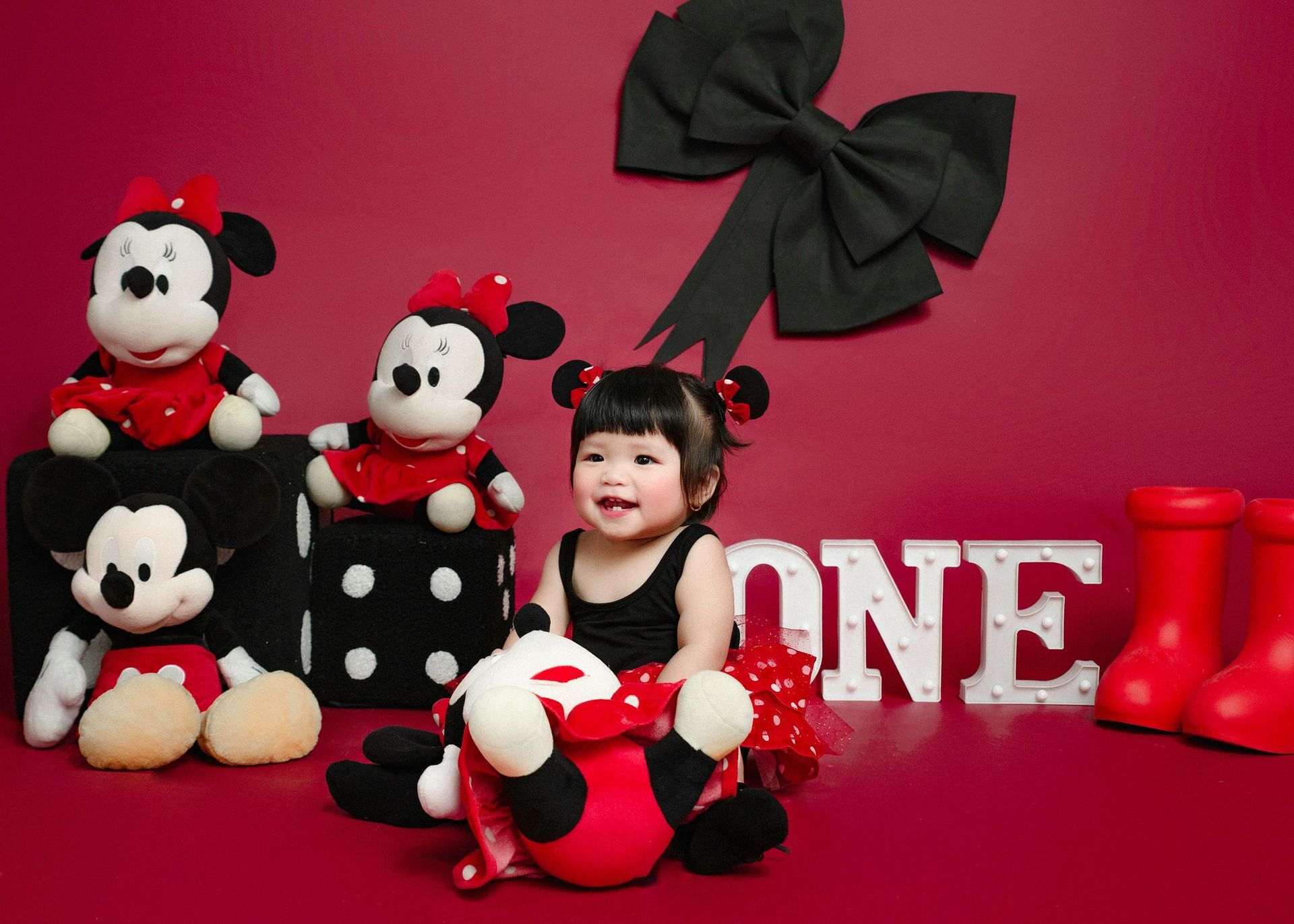 Smiling baby with Mickey and Minnie Mouse plushies, red backdrop, 
