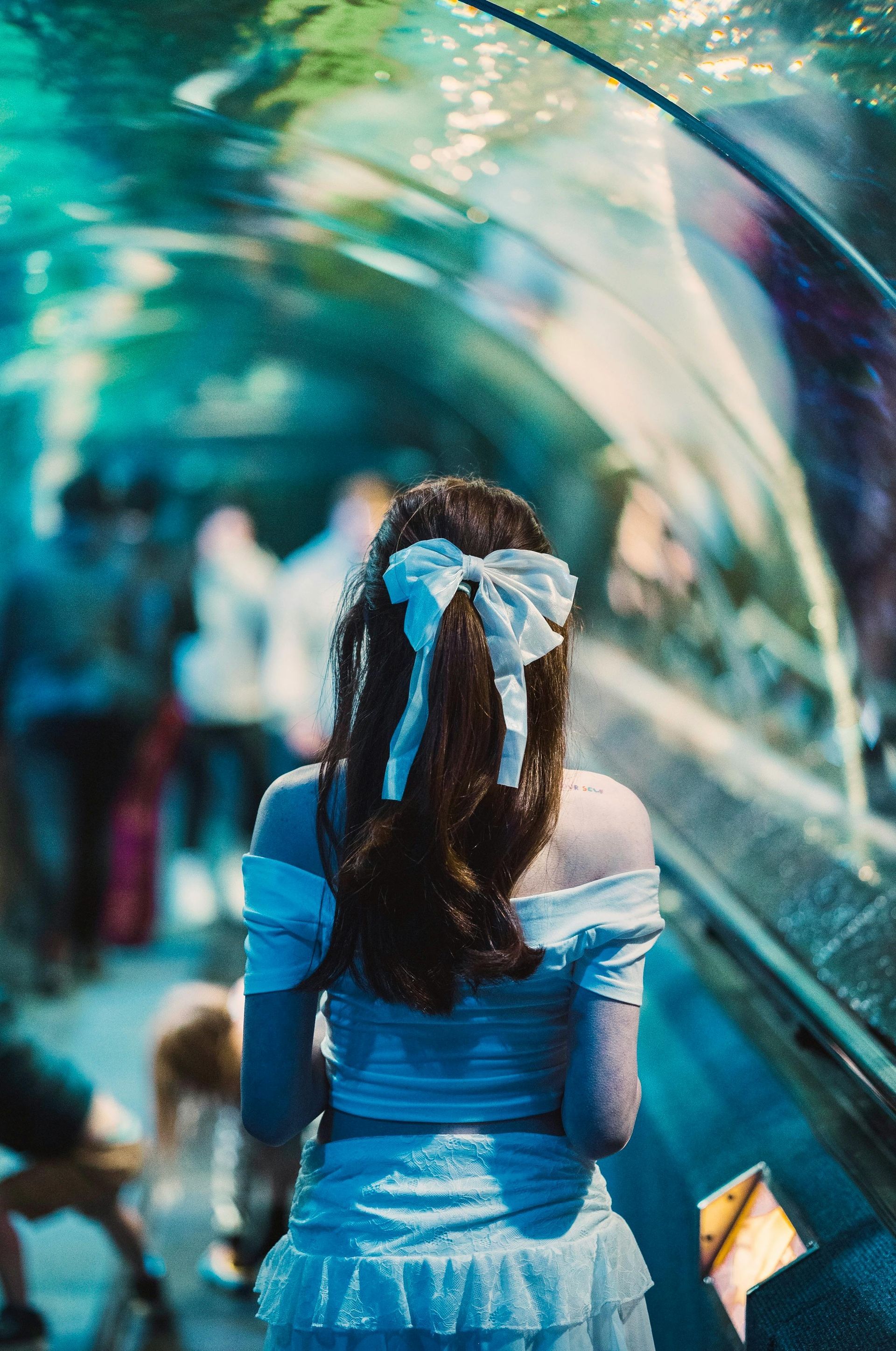 Woman with a white bow in her hair looks into an aquarium tunnel, wearing a blue off-the-shoulder top.