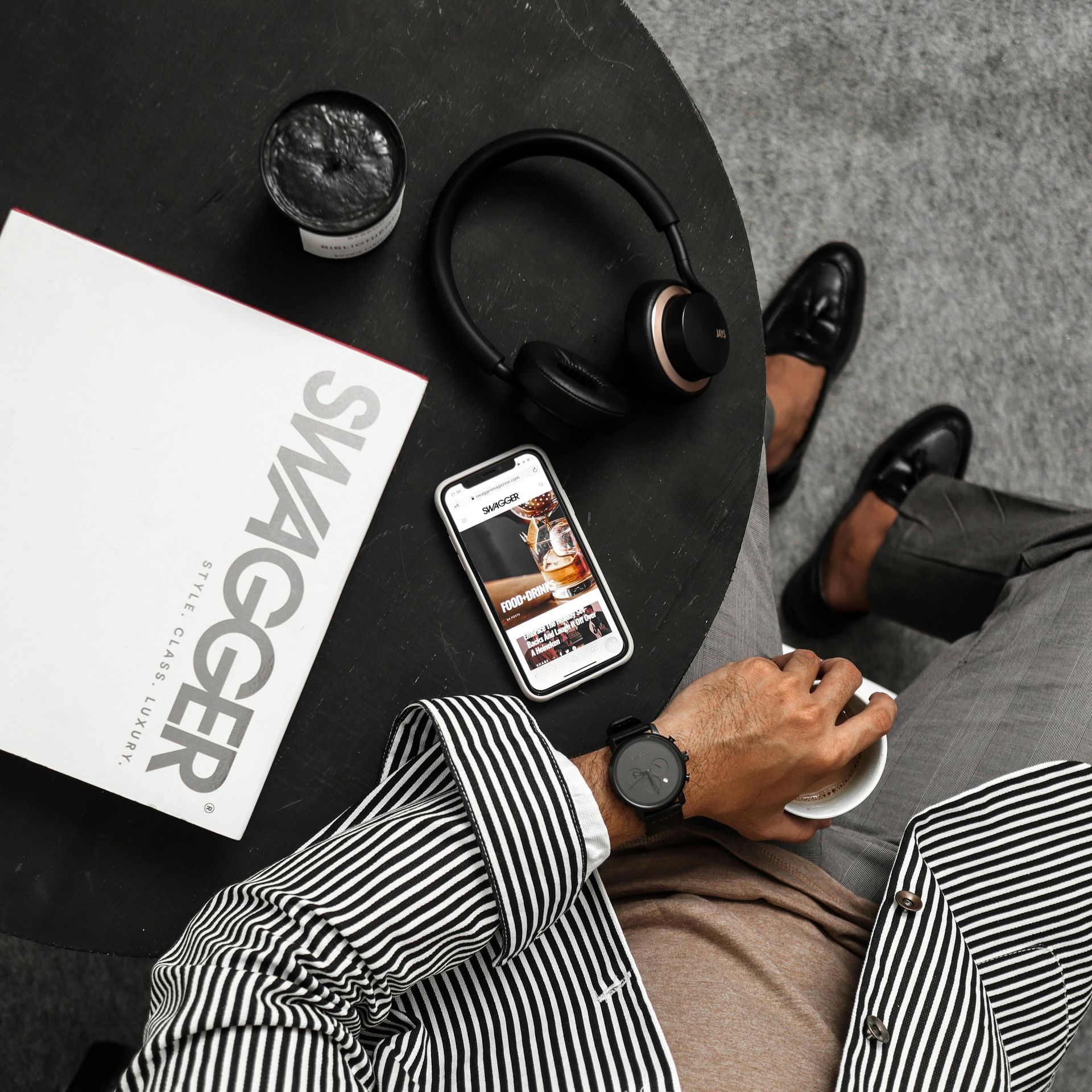Person in striped jacket with watch, holding coffee, phone, headphones, magazine, shoes on table.
