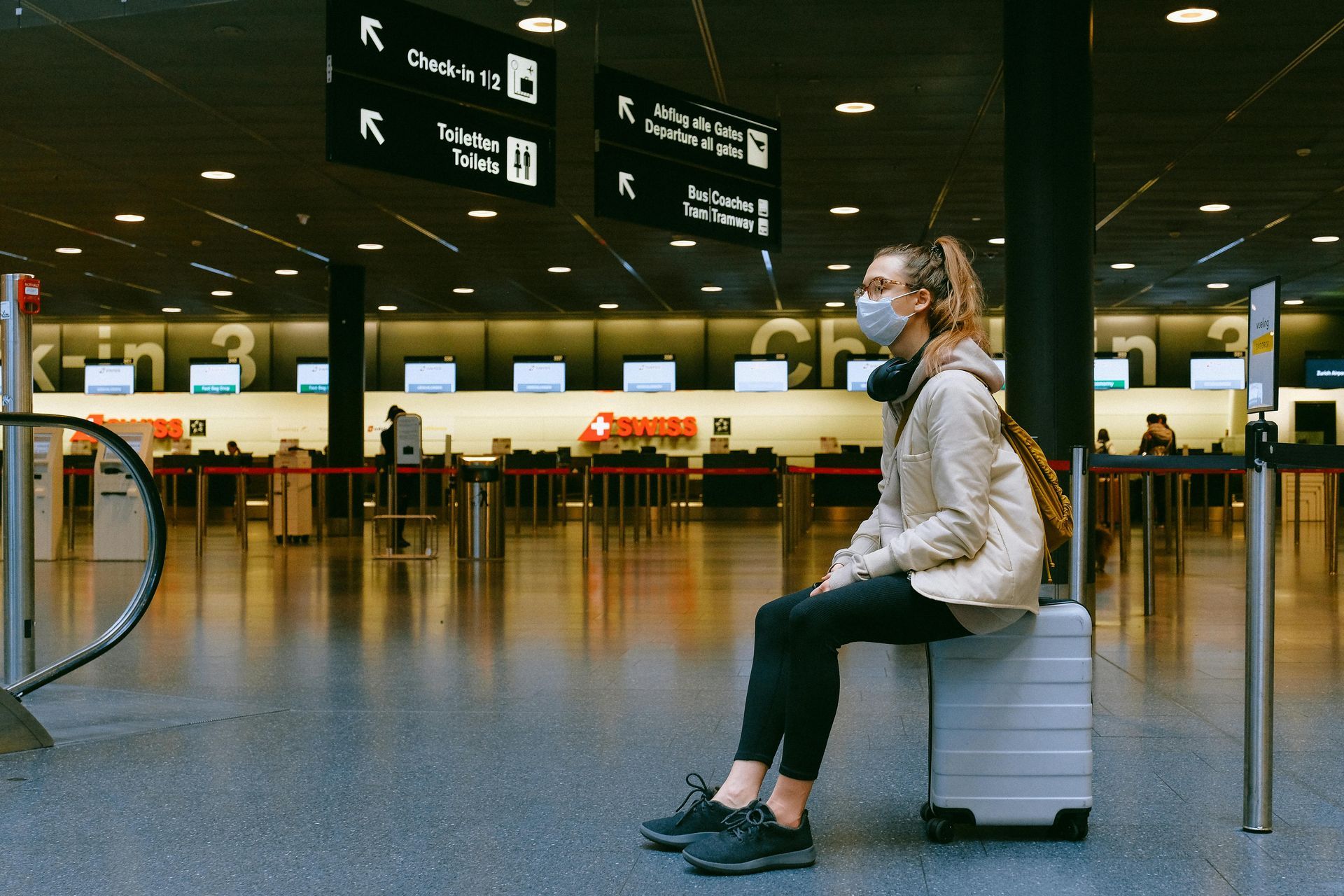 Woman wearing a face mask sits on a suitcase in an empty airport terminal, waiting.