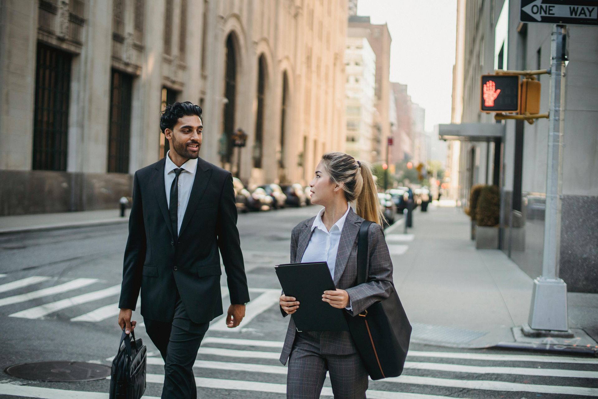 Two business people crossing a street, man carrying briefcase, woman holding tablet. Buildings in background.
