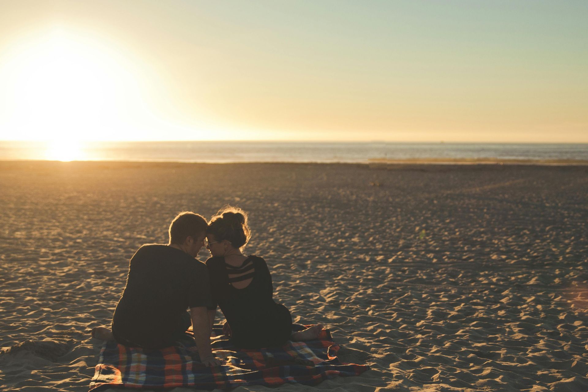 Couple on a blanket at beach, silhouetted against a golden sunset over the ocean.