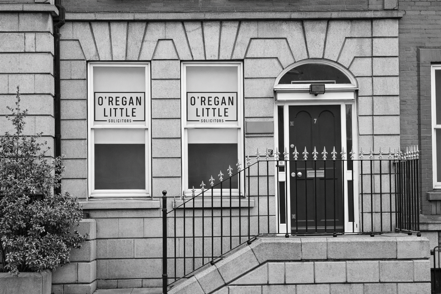 Gray stone building with two windows, a dark arched door, and a wrought-iron railing.