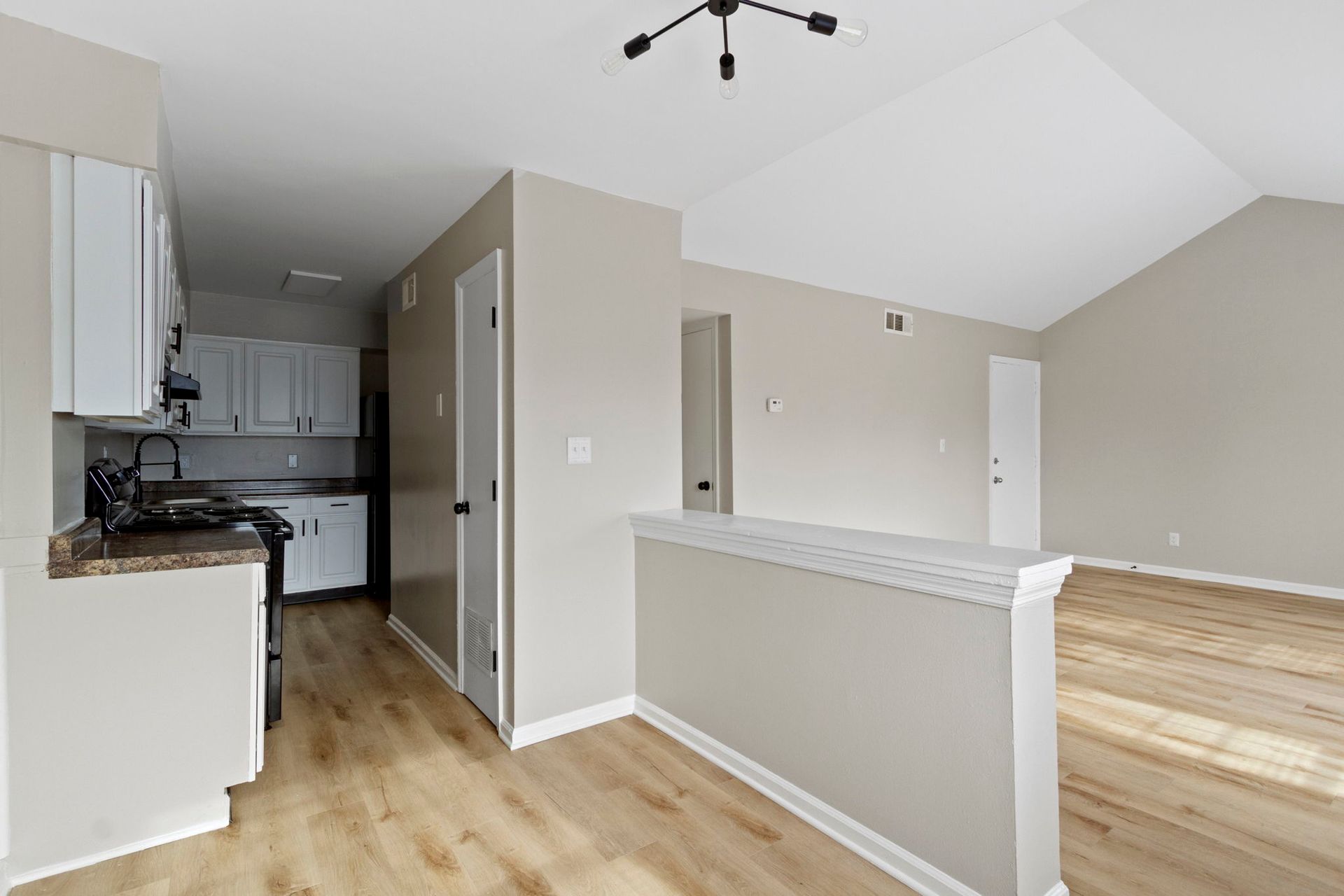 Open-plan apartment with kitchen, living area, and a half wall. Light-colored walls, white cabinets, and wood-look flooring.