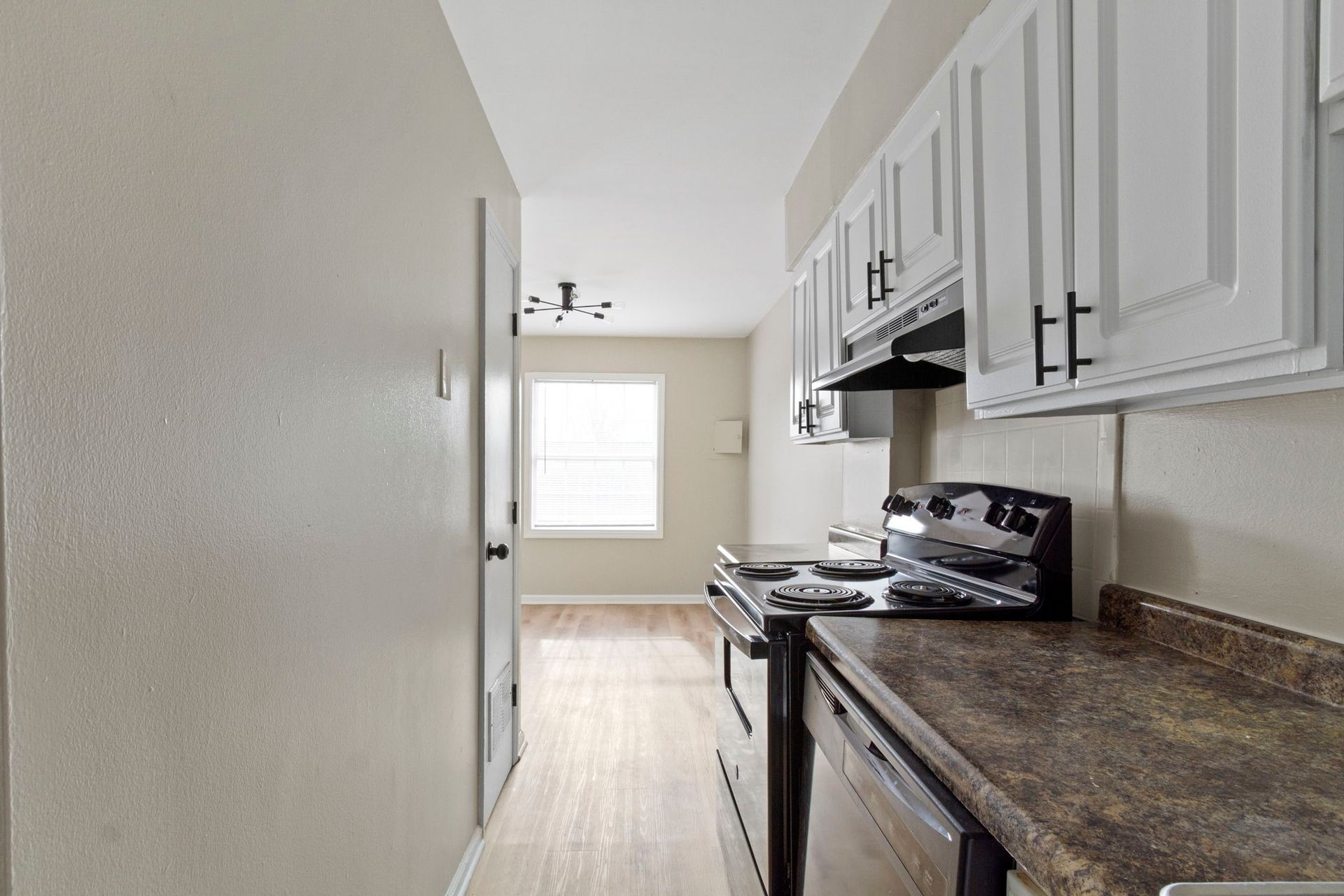 Narrow kitchen with white cabinets, black appliances, and a window at the end of the hallway.