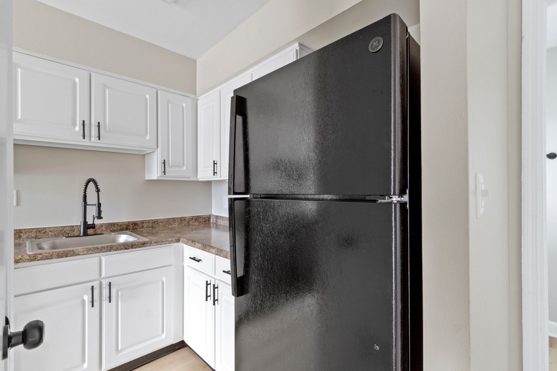 Black refrigerator next to white cabinets in a small kitchen with a sink and black faucet.
