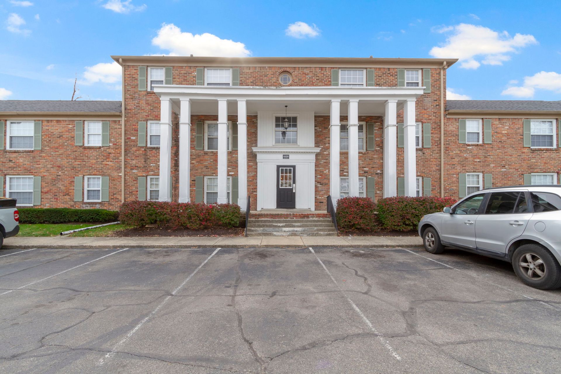 Apartment building with white columns, red brick, gray car parked in front, overcast sky.