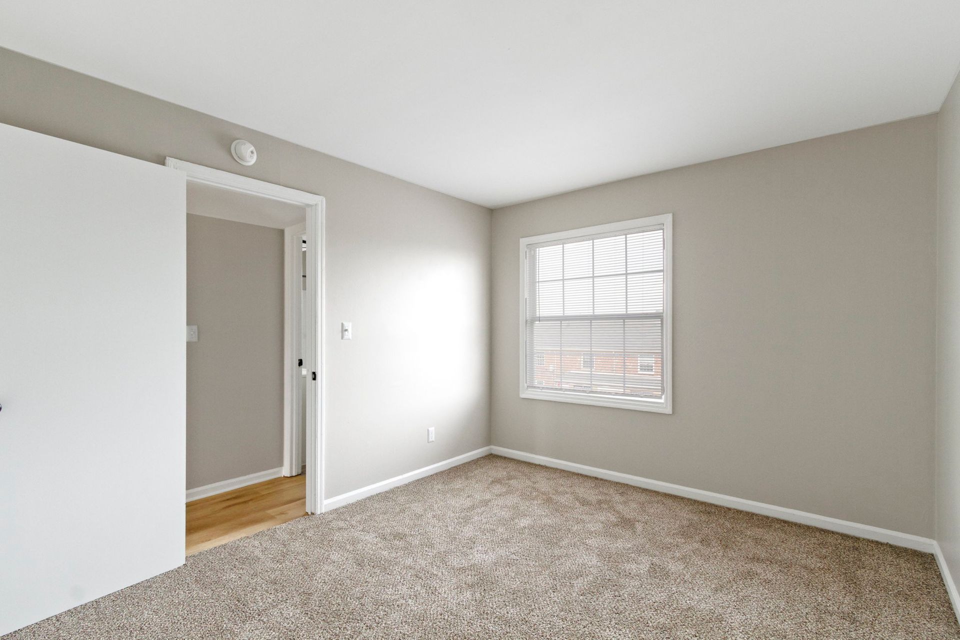 Empty bedroom with neutral walls, carpet, and window with blinds; a partially open doorway.