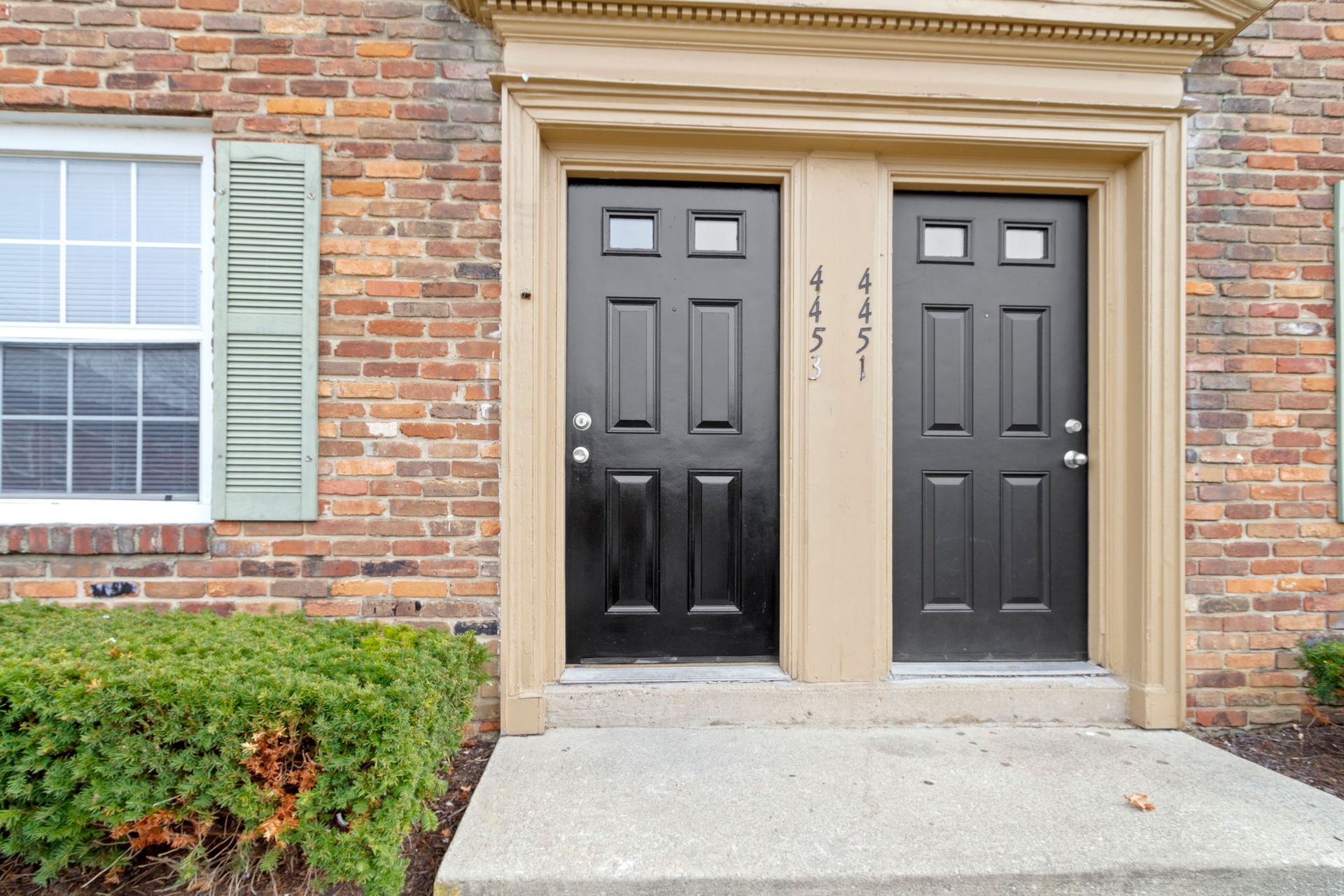 Two black front doors with address numbers, tan trim, brick building, and bushes.